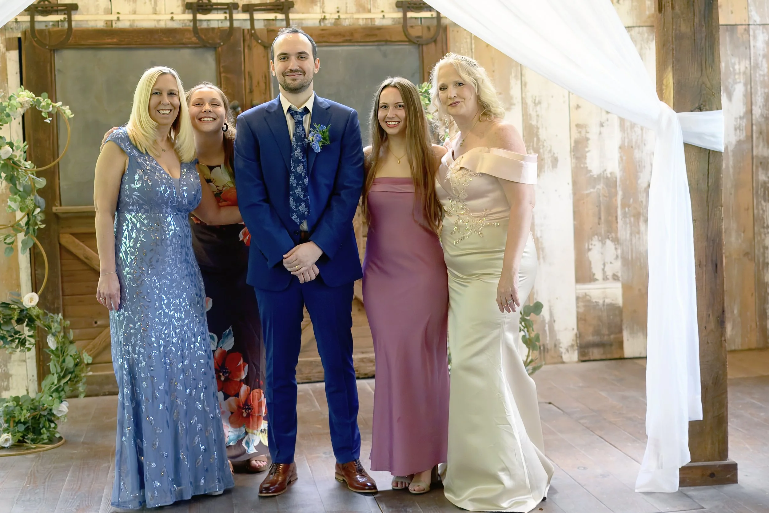 Group of five people standing together inside a rustic venue during a celebration, dressed in formal and semi-formal attire, smiling at the camera.