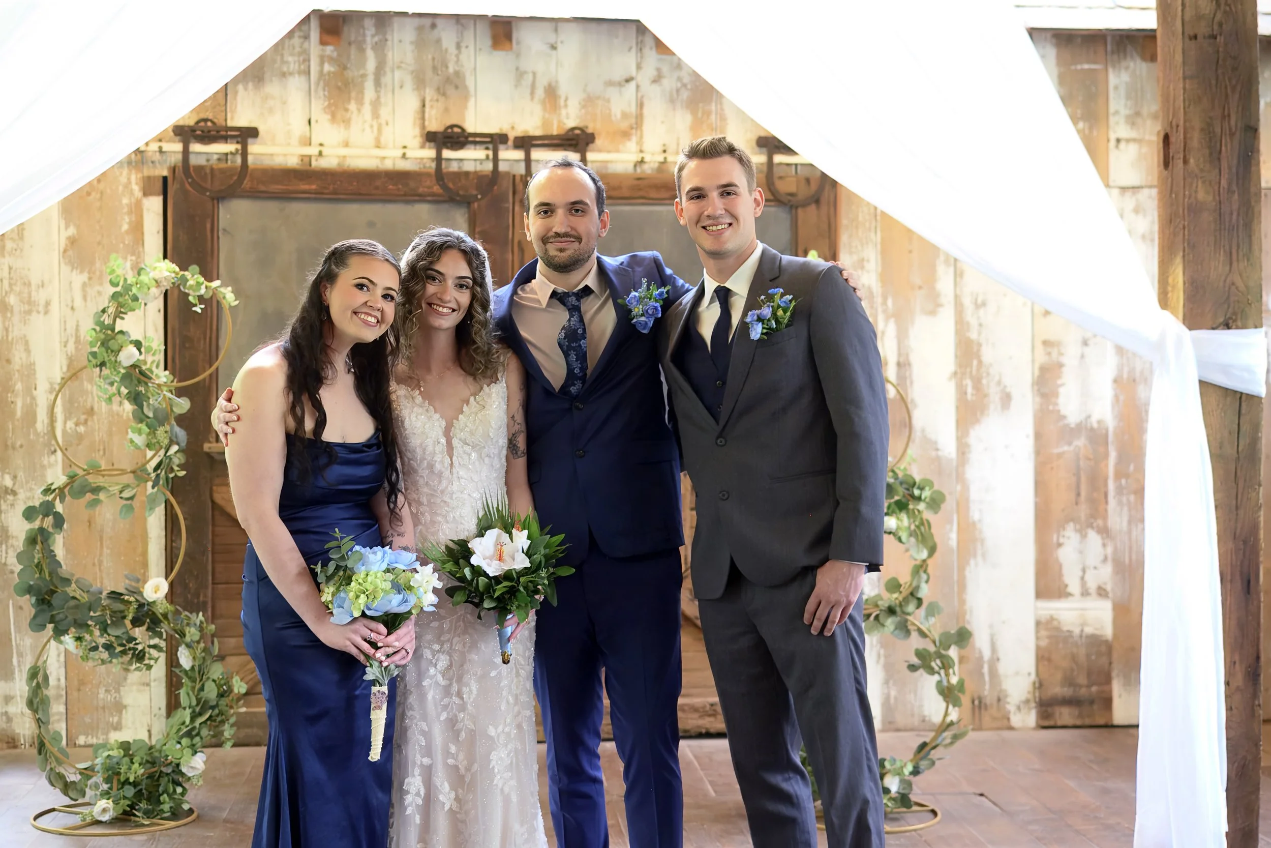 Group of four people, including a bride, a groom, and two women, standing in a wedding venue with rustic wooden walls and floral decorations.