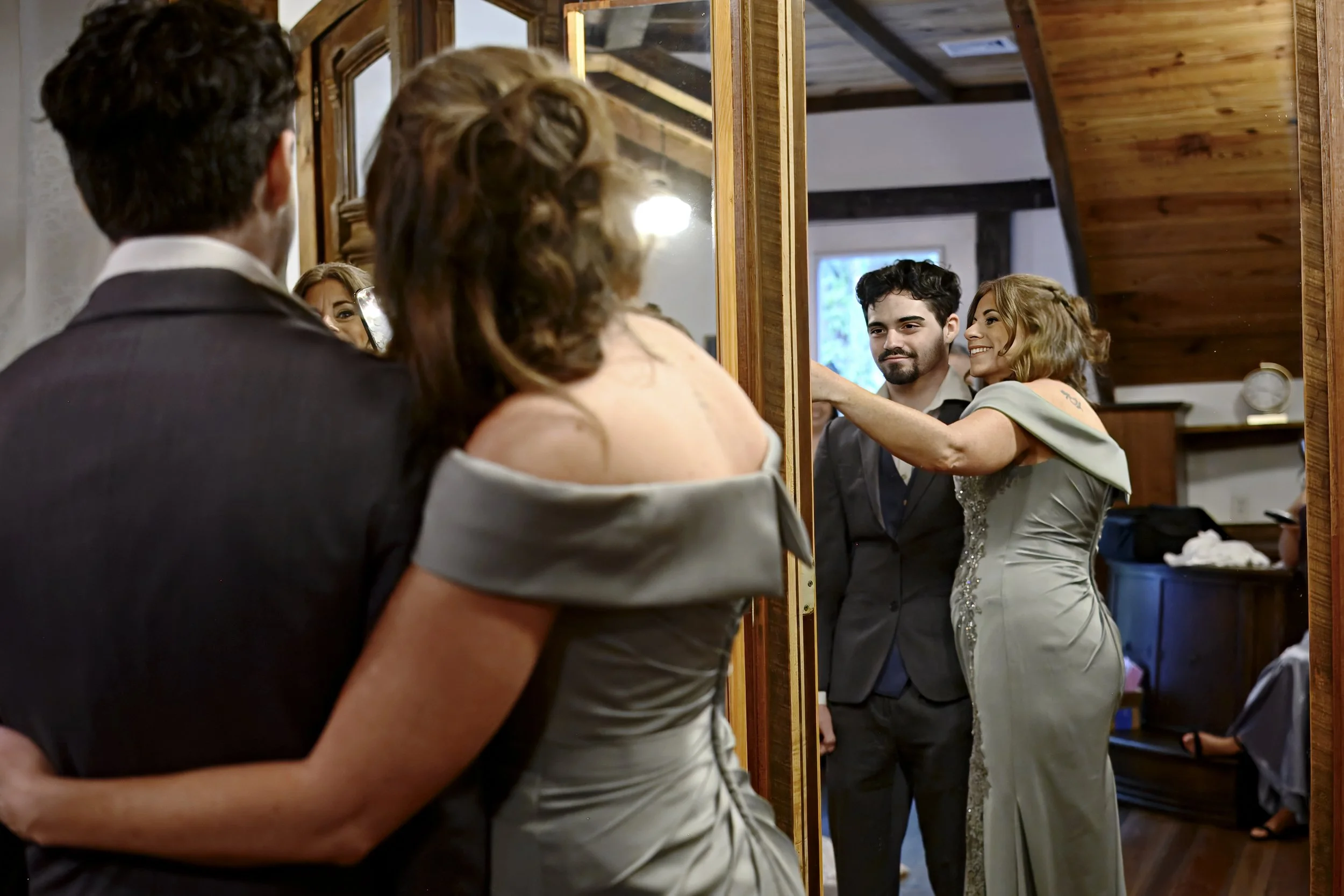 A woman and a man are looking at themselves in a mirror. They appear to be at a wedding or formal event, with the woman in a dress and the man in a suit. They are smiling and reaching out to each other.