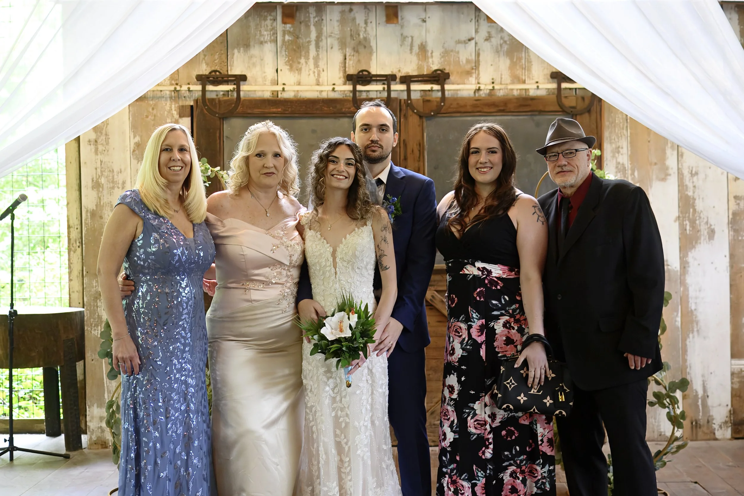 Group of six people, including a bride and groom, posing for a photo at a wedding venue with rustic decor and floral arrangements.