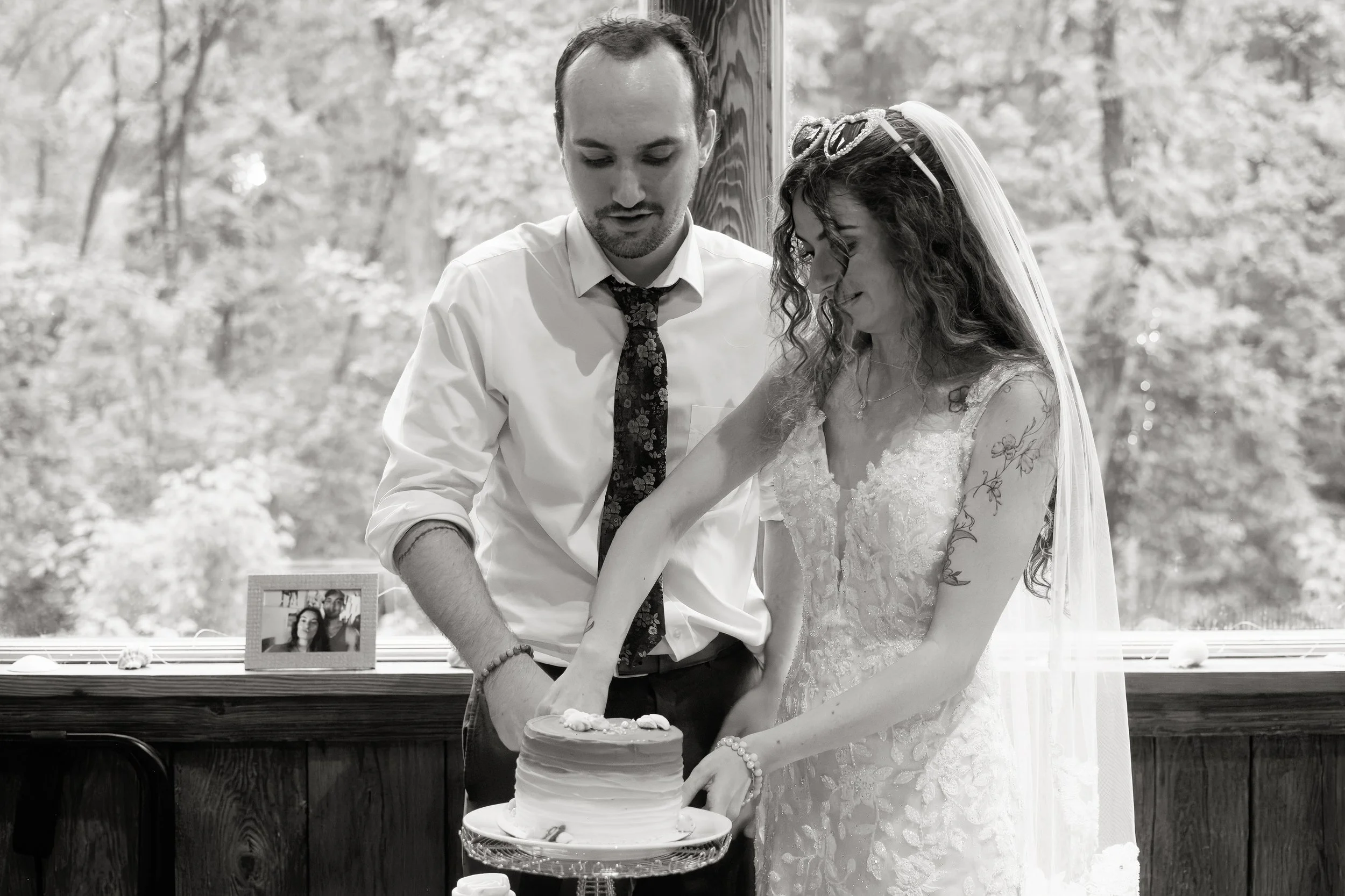 A groom and bride are cutting a wedding cake together during their wedding reception. The bride is wearing a wedding dress with lace details and a veil, while the groom is wearing a white shirt and a floral tie. A framed photo is placed on the table 