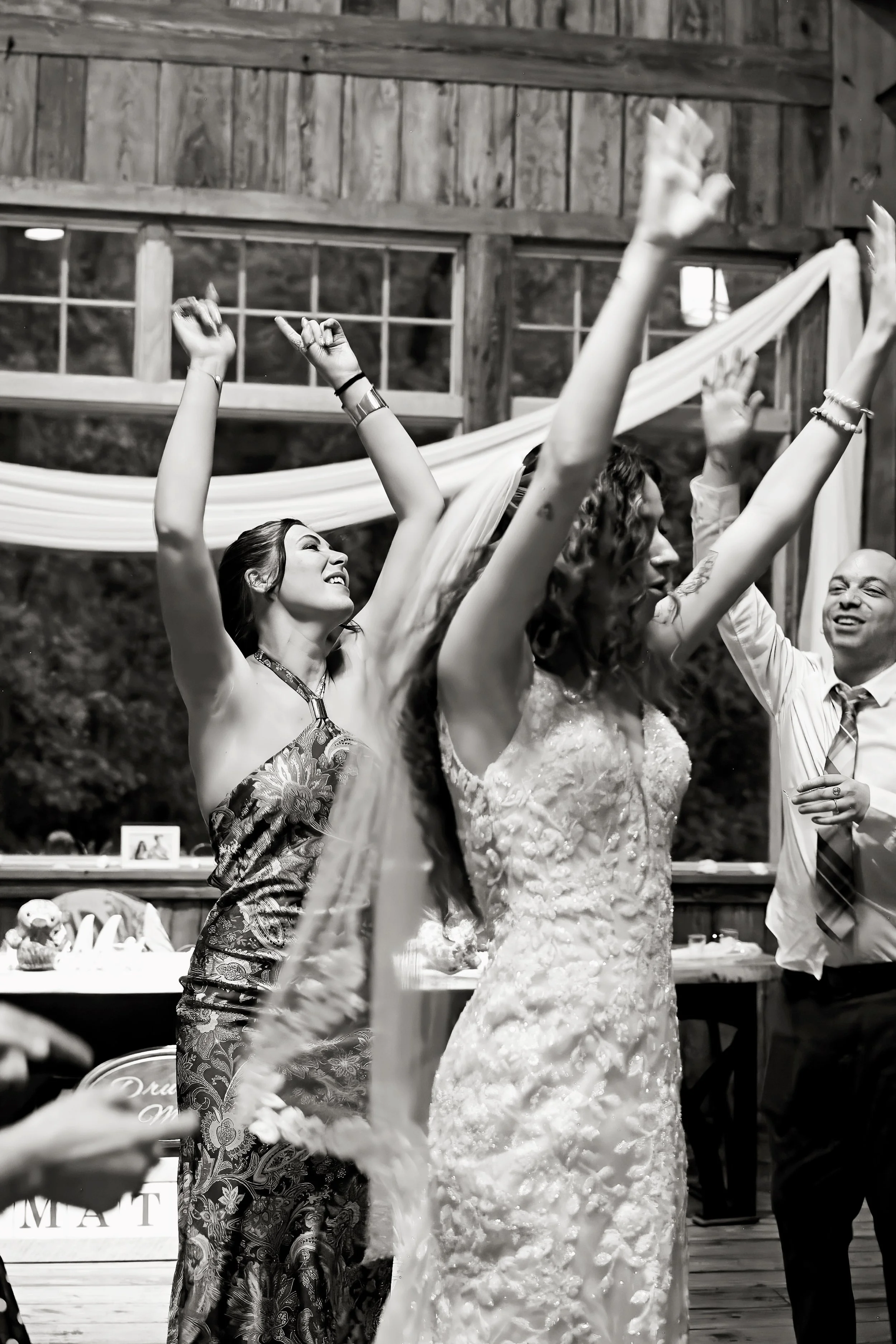 People celebrating at a wedding reception inside a wooden venue. A woman in a floral dress smiles with her arm raised, while other guests also raise their hands in celebration.