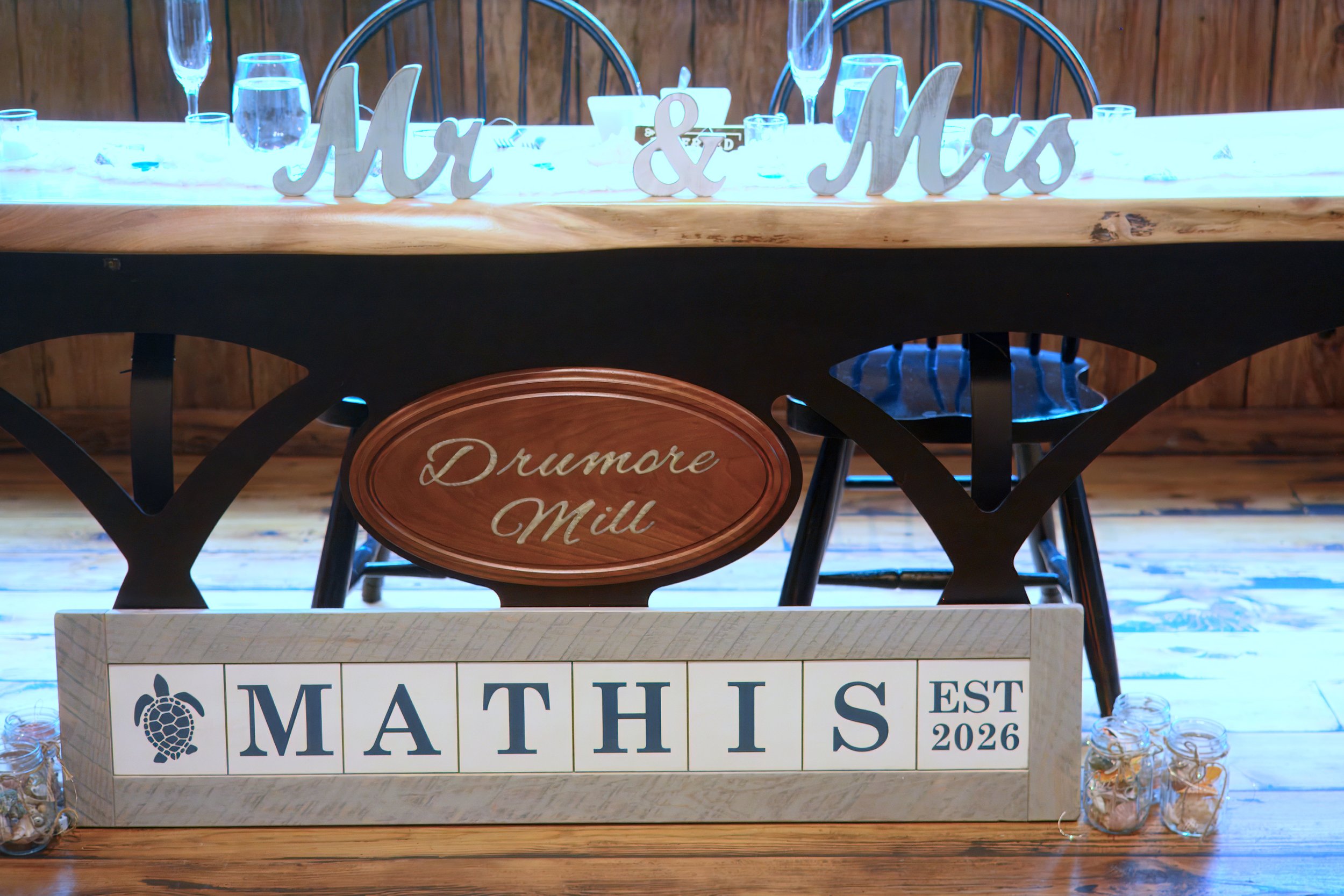 A wedding reception table decorated for Mother's Day with a wooden sign that reads "Mom & Mums" in white cursive letters. The table features glasses of water, champagne flutes, small jars filled with candies, and a sign that says "Mathis Est 2026." T