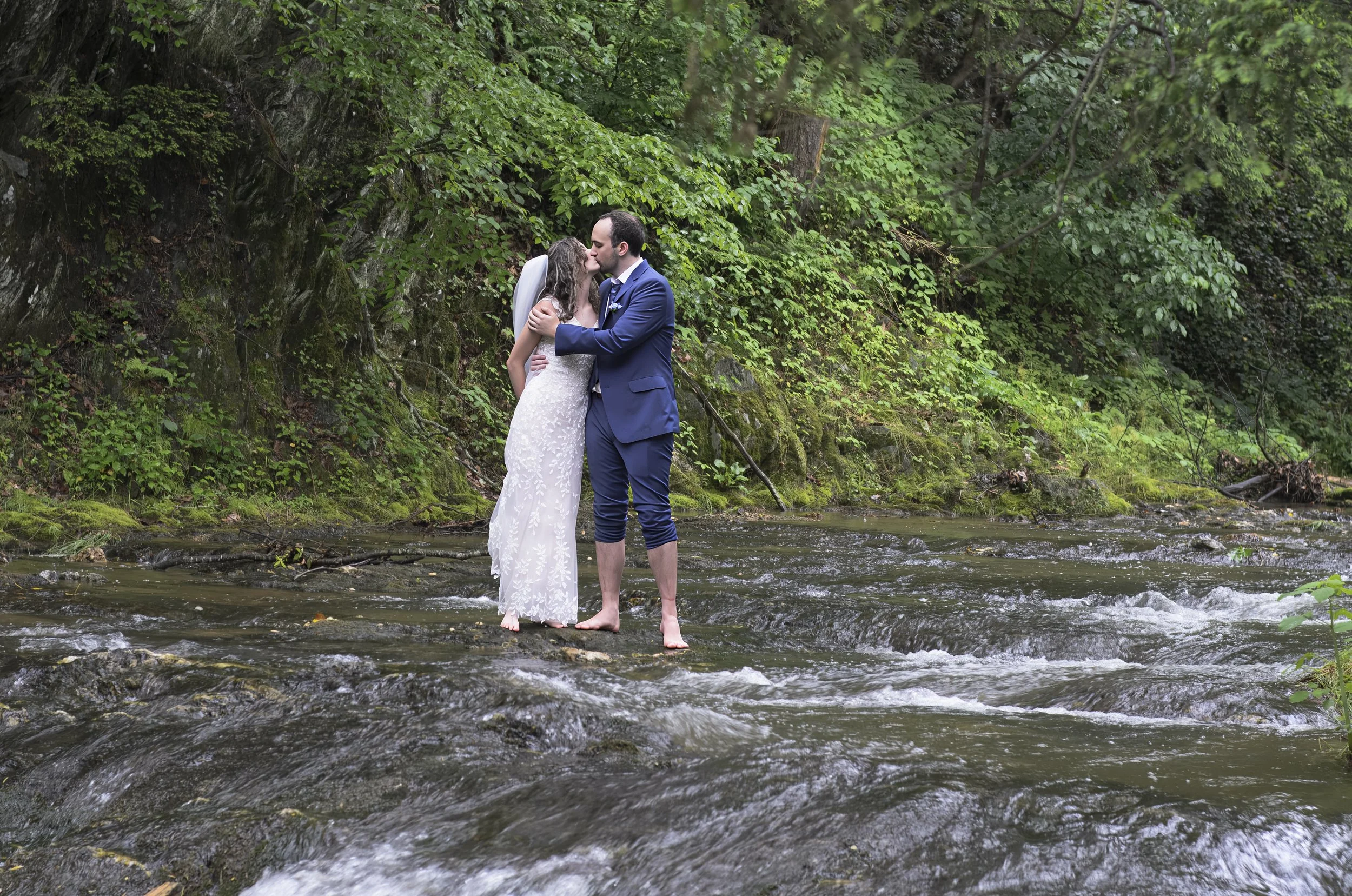 A couple dressed in wedding attire kisses and embraces in the middle of a stream, with lush green foliage in the background.