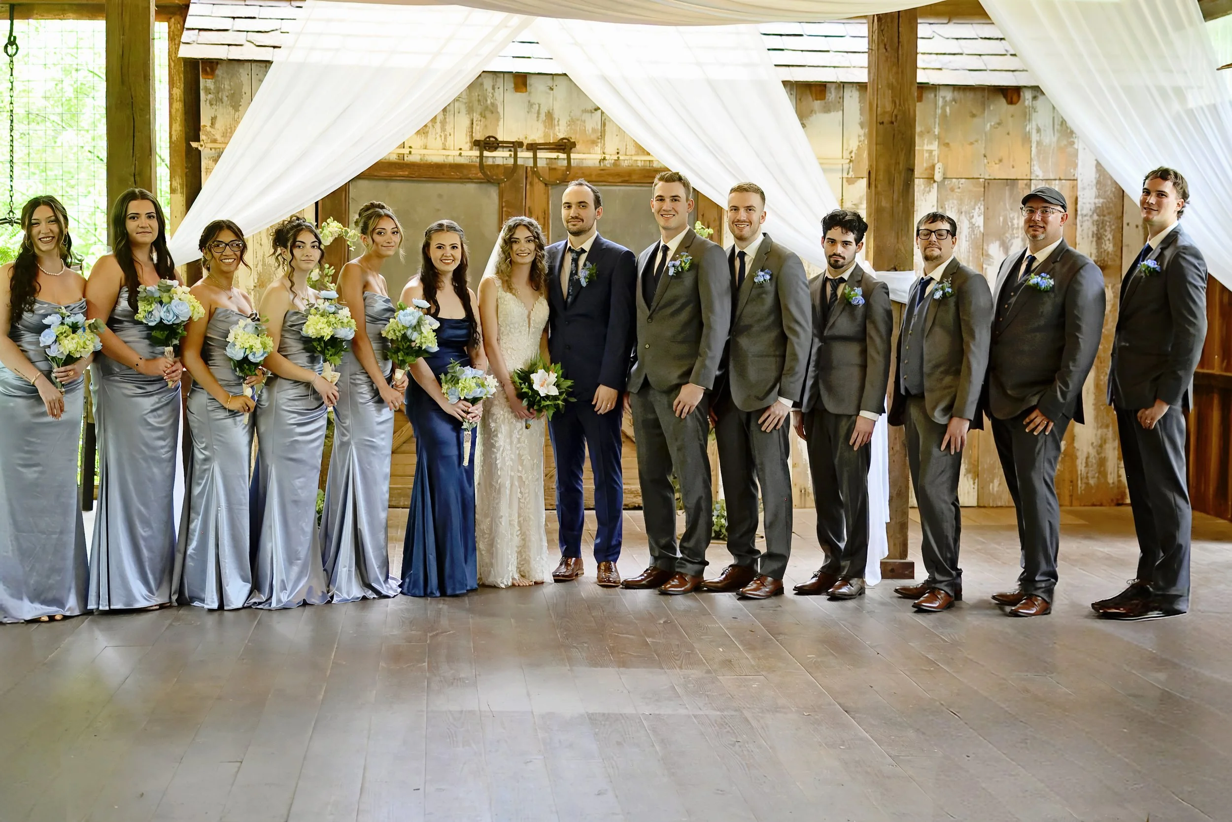 A group of wedding guests, including bridesmaids and groomsmen, standing in a rustic venue decorated with white drapery and flowers, posing for a photo.