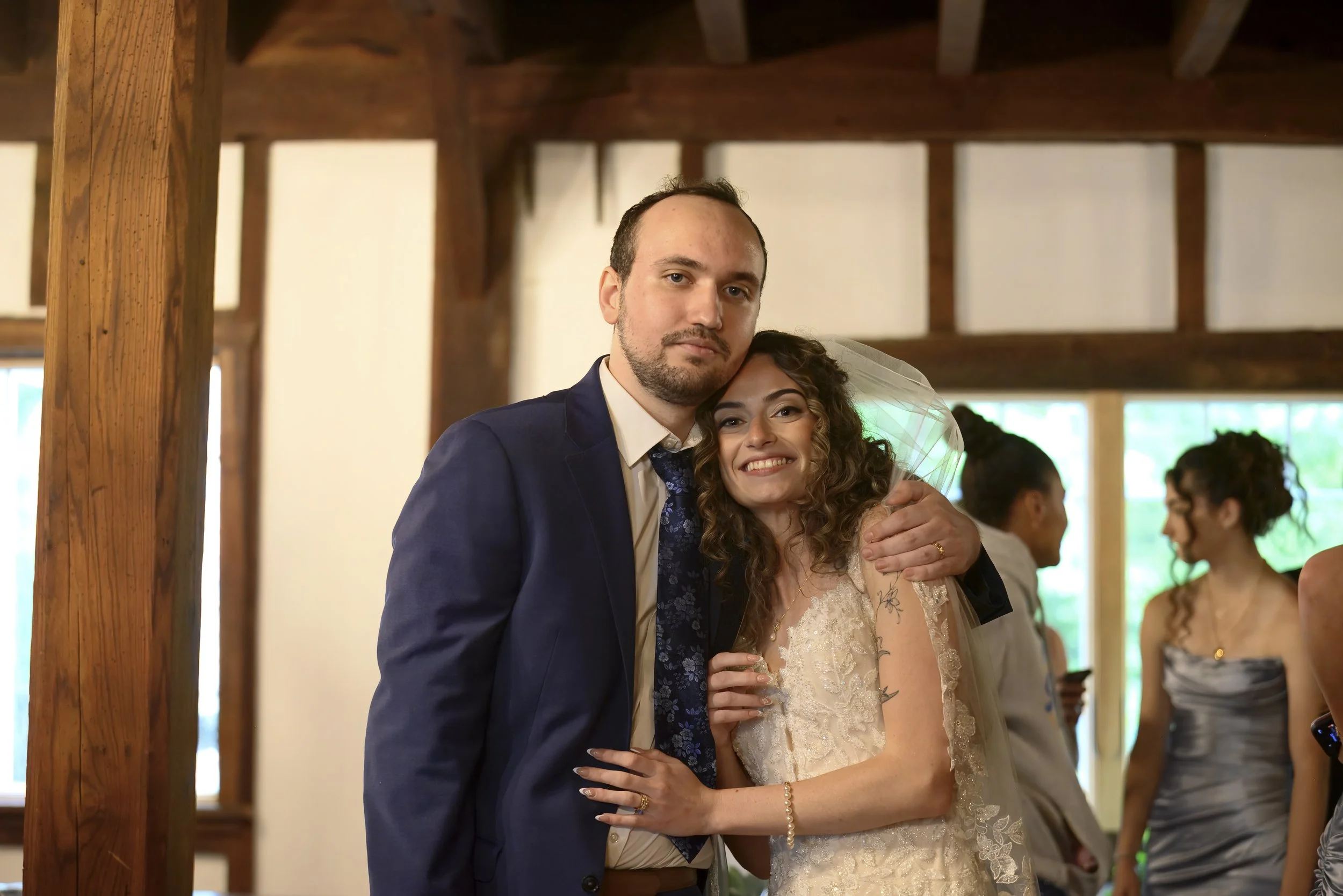 A man and woman embracing at a wedding reception. The man is wearing a navy blue suit, and the woman is in a lace wedding dress with a veil. They are indoors with wooden beams and other guests in the background.