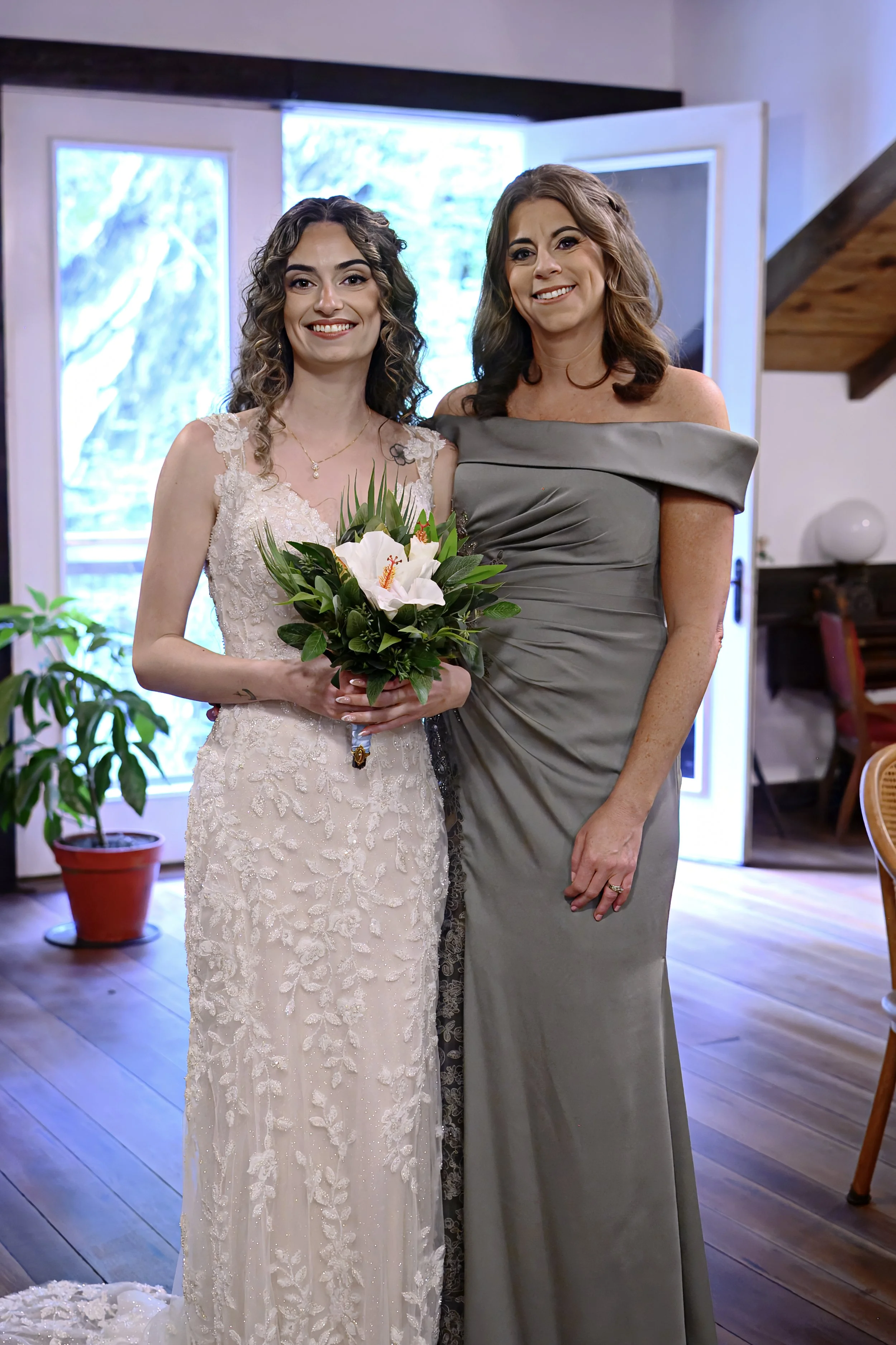 Two women standing together indoors, one wearing a wedding dress holding a bouquet and the other in a gray off-shoulder dress, smiling at the camera with open windows in the background.