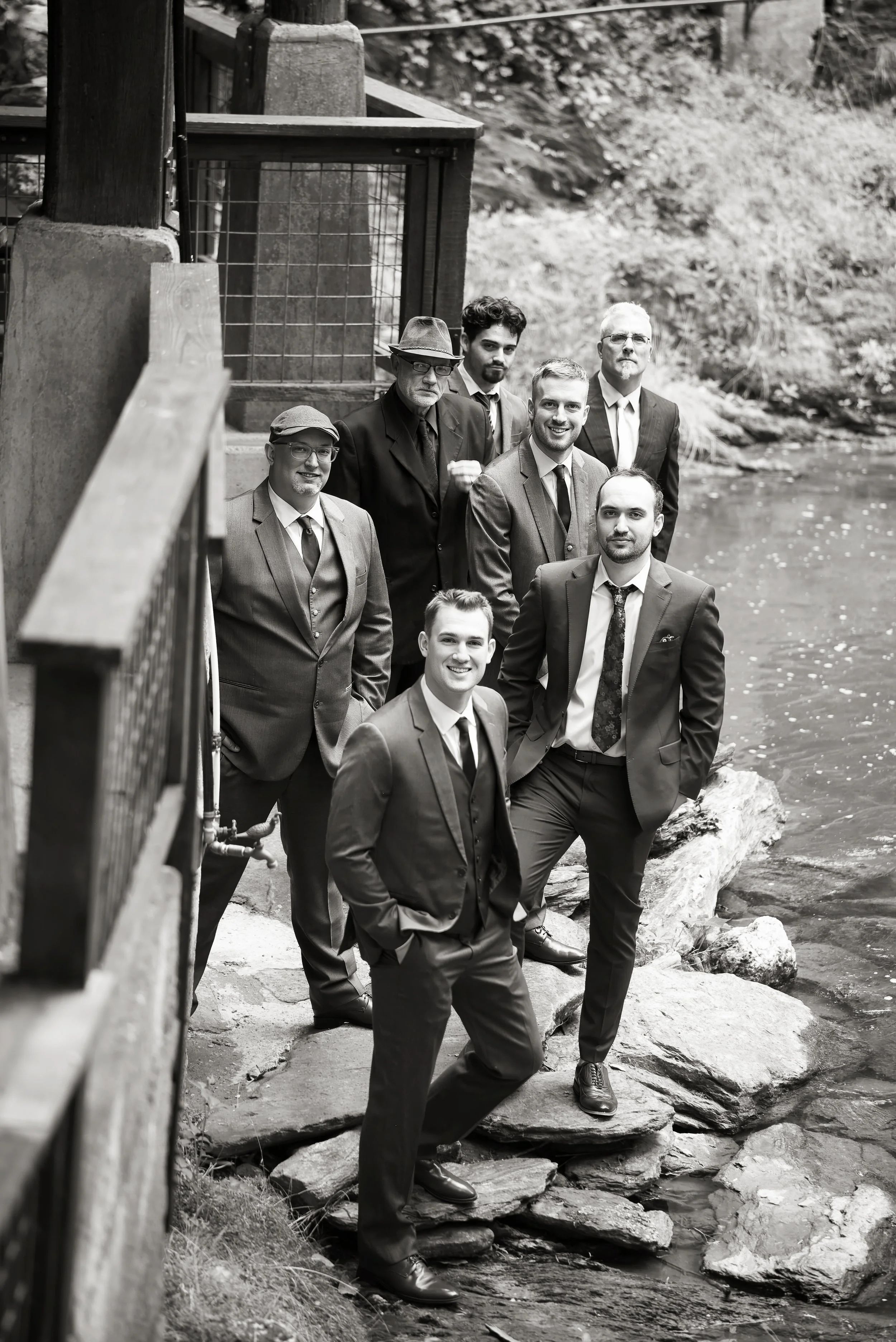 Black and white photo of six men in suits standing on rocks near a river, with a wooden fence and lush greenery in the background.