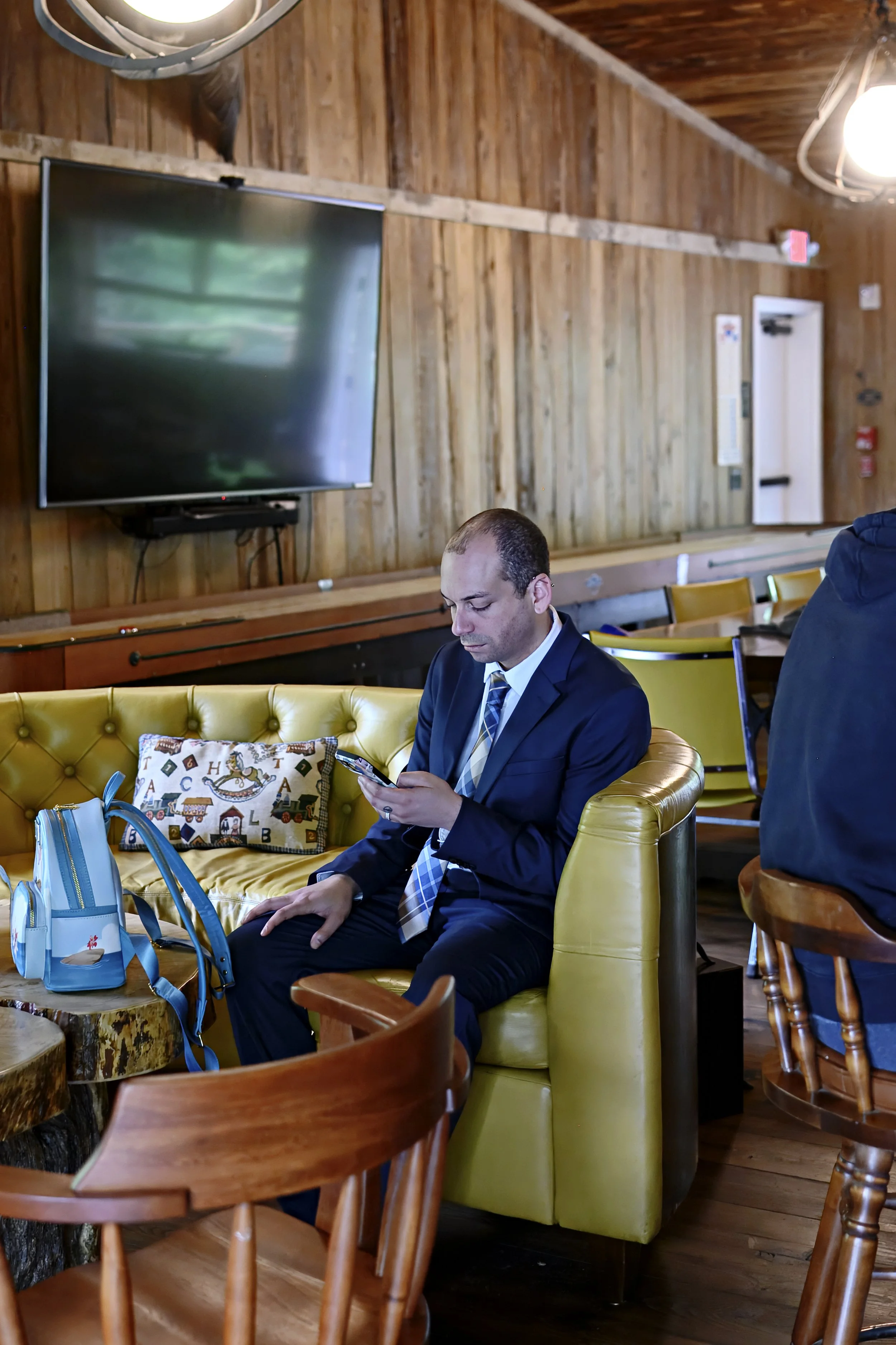 A man in a business suit sitting on a yellow sofa, looking at his phone, with a colorful decorative pillow beside him in a wooden interior space.