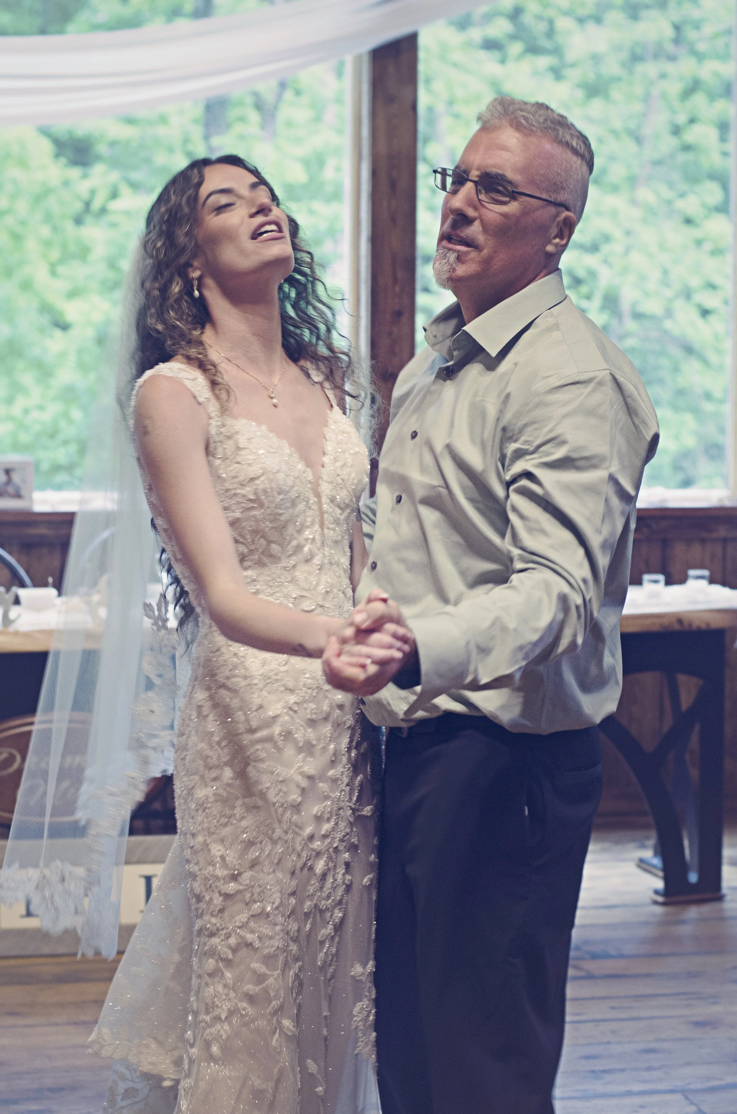 A woman in a wedding dress and a man in a dress shirt are dancing at a wedding reception indoors with large windows showing greenery outside.