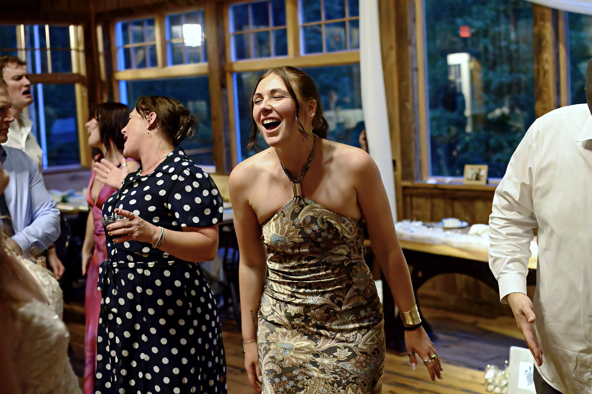 A group of people at a social gathering inside a wooden building, with windows revealing trees outside. One woman with short hair, wearing a patterned dress and jewelry, is laughing and walking among others.
