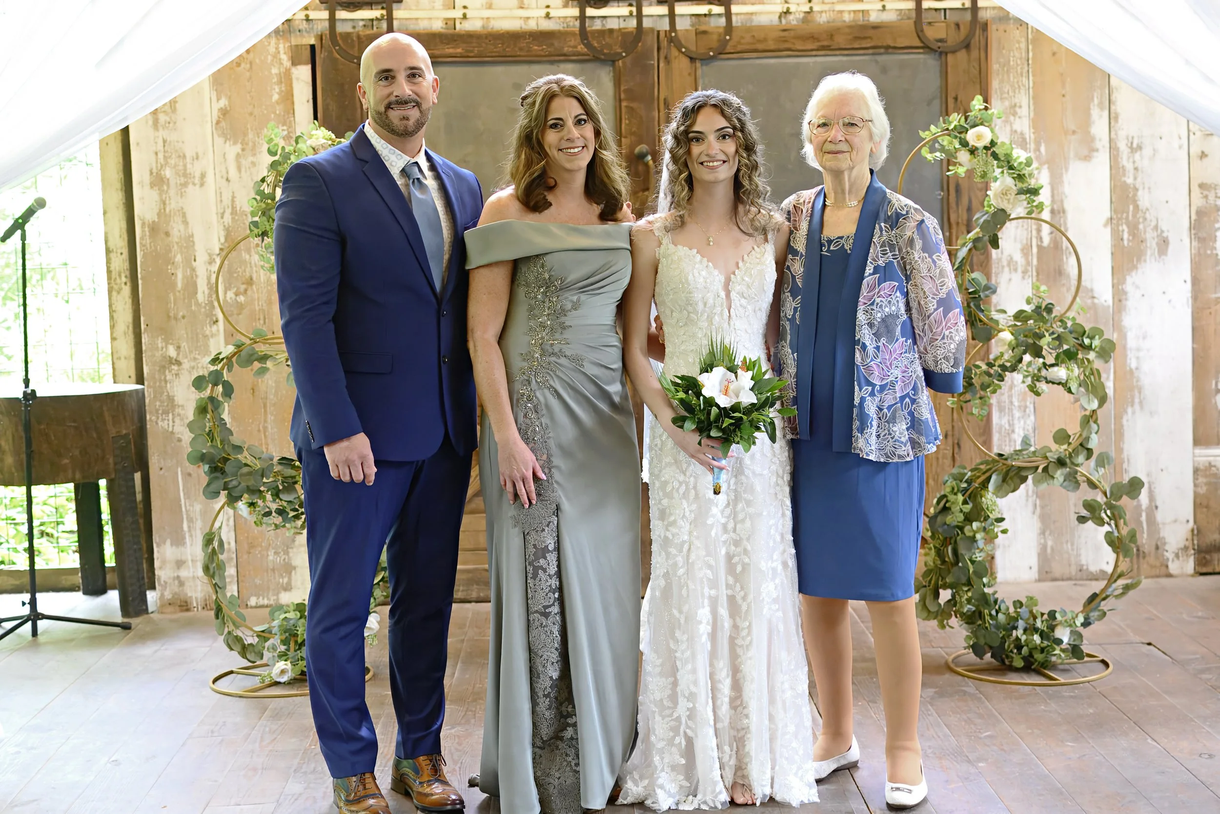 Group of five individuals at a wedding ceremony, standing together in front of a rustic wooden backdrop decorated with greenery and flowers, smiling at the camera.