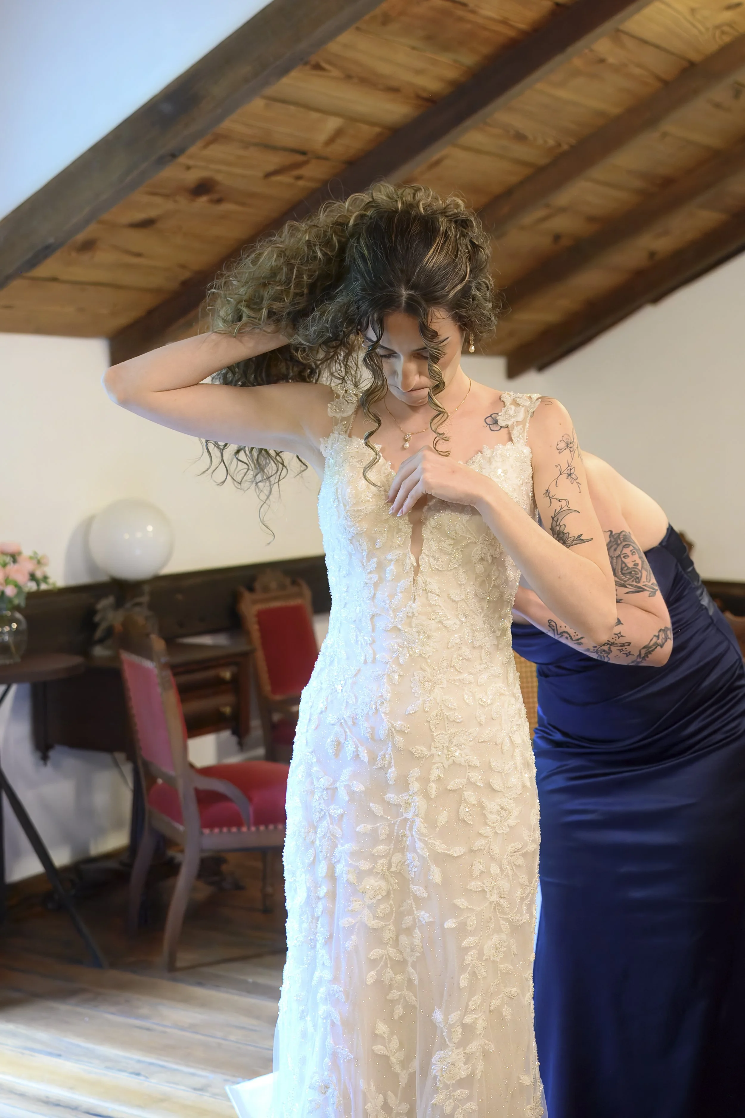 A woman in a white wedding dress with lace details, standing indoors with an arched wooden ceiling above her. She has curly hair and appears to be adjusting her dress or hair. Another person in a dark dress is assisting her. The background features c
