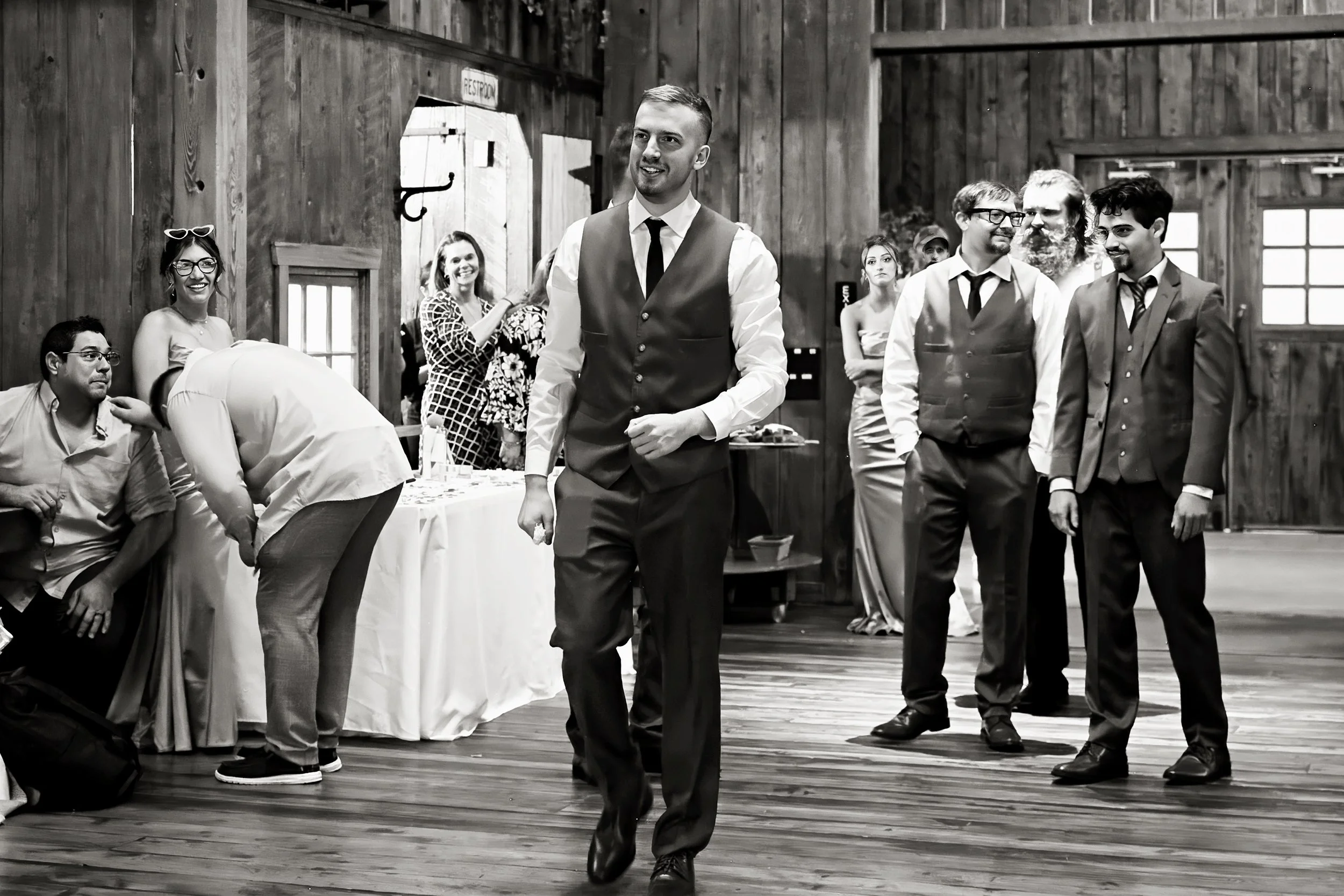 A man in a vest and tie walking in a rustic wooden wedding venue with guests smiling and watching him.