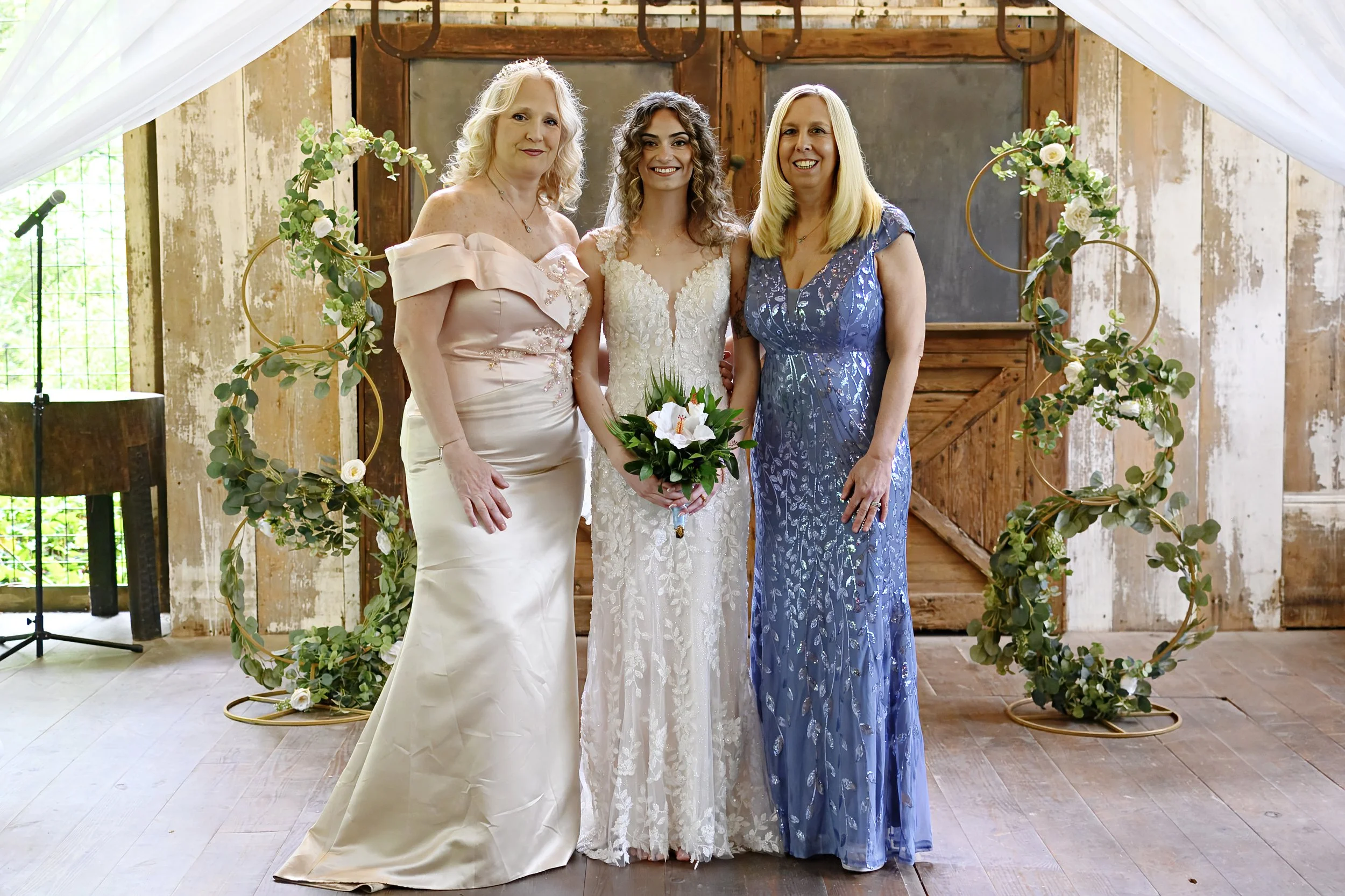 A bride in a white lace wedding dress holding a bouquet, standing with two women, one in an off-the-shoulder light pink gown and the other in a blue floral dress, in front of a decorative circular floral arch at an indoor wedding ceremony.