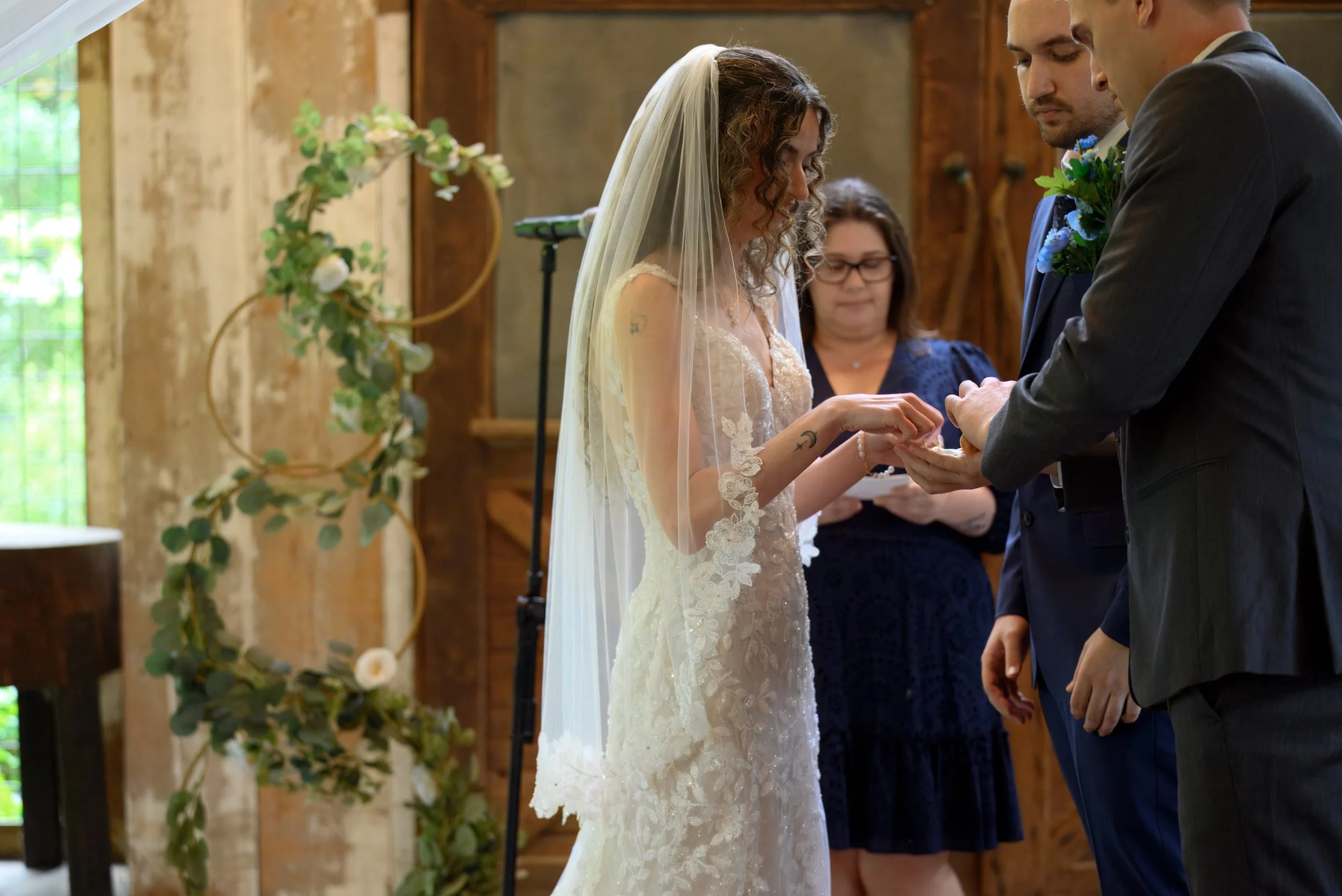 A bride in a lace wedding dress and veil and a groom in a dark suit exchange rings during a wedding ceremony in a rustic indoor setting with wooden walls and decorated arch.