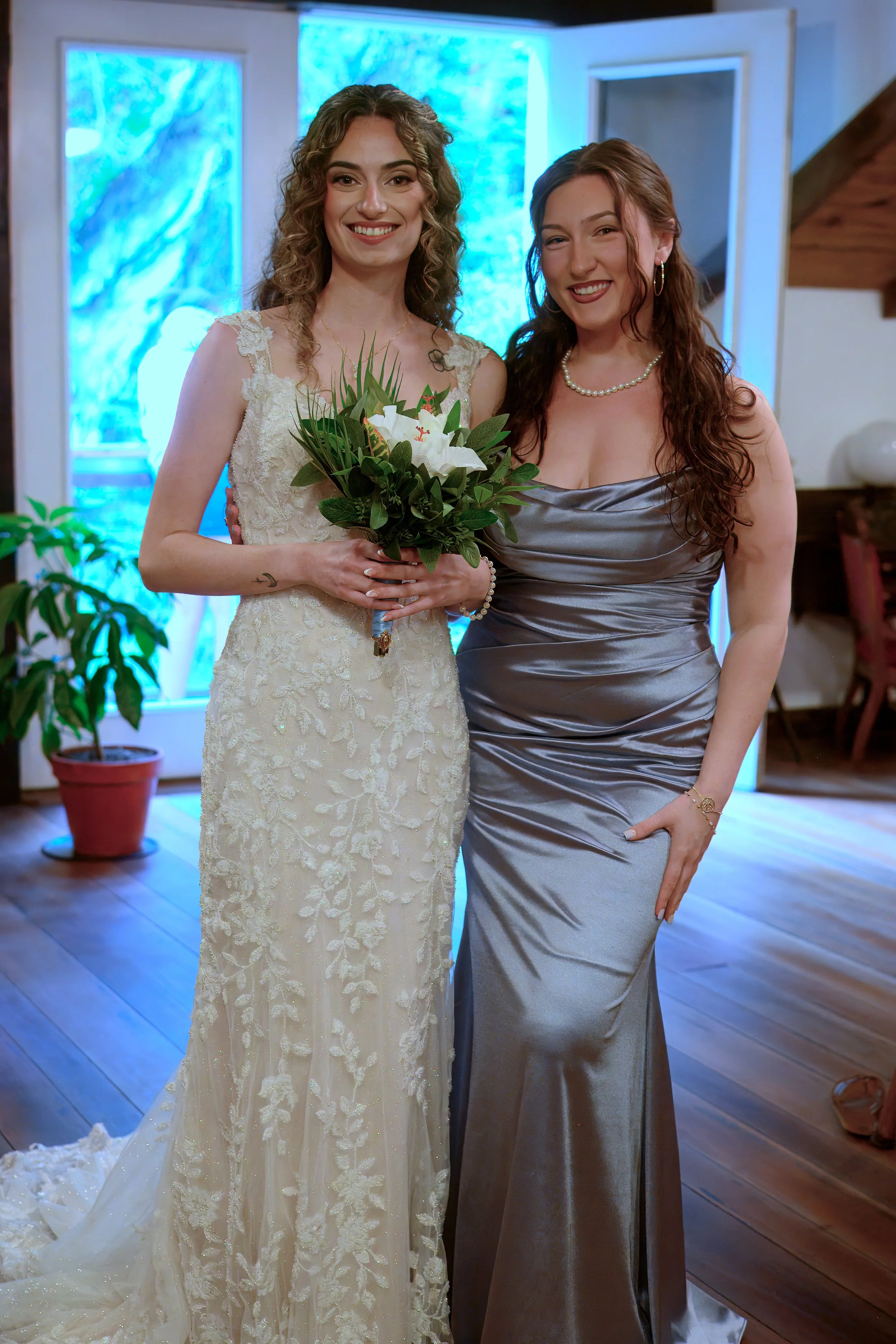 Two women dressed in formal attire, one in a white wedding dress holding a bouquet, the other in a silver gown with a pearl necklace, standing inside a decorated room with blue lighting in the background.