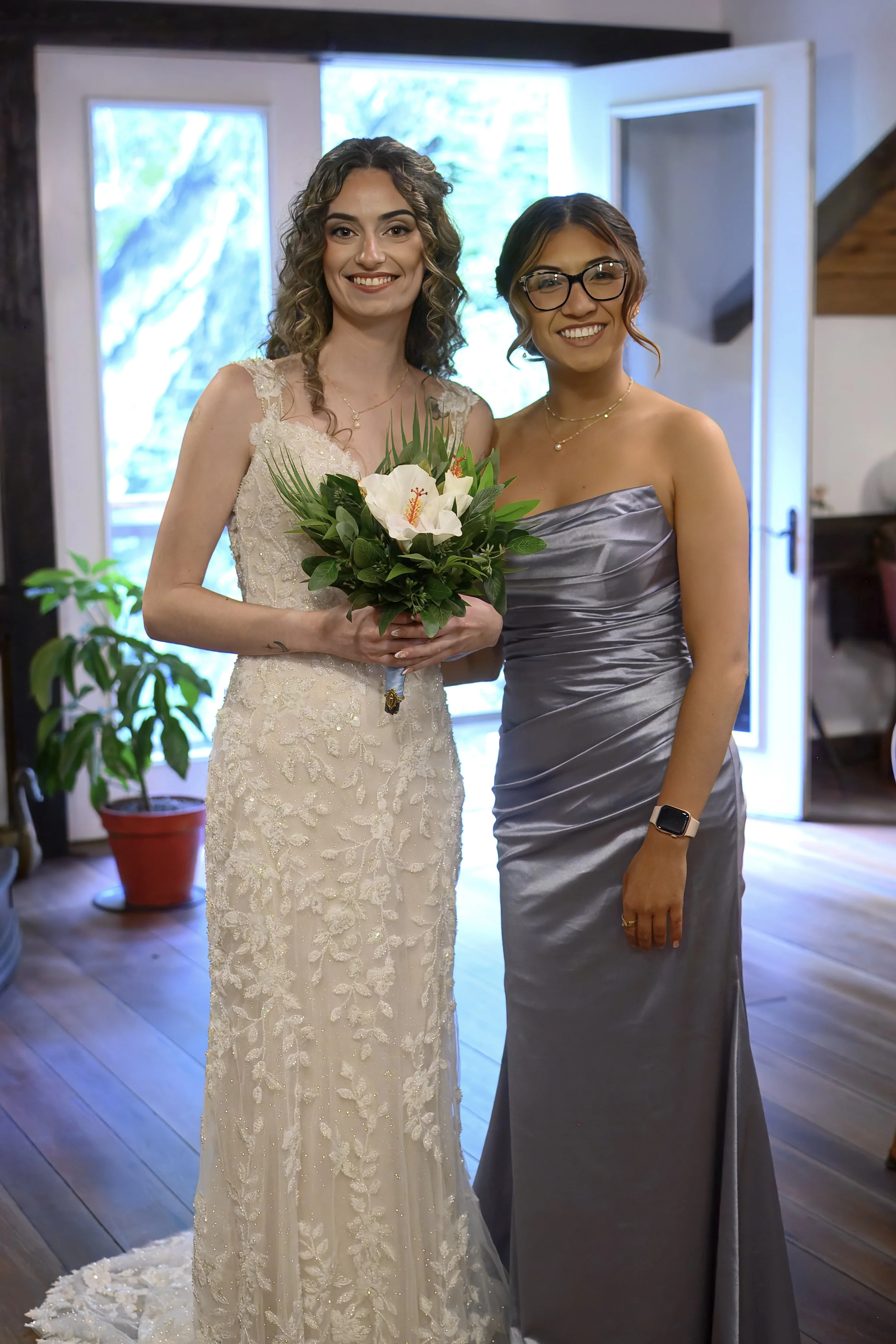 Two women standing inside a house near a door, holding a bouquet of white flowers. One woman is wearing a white lace wedding dress, and the other is wearing a strapless, shiny silver dress with glasses and jewelry. There are large windows and potted 