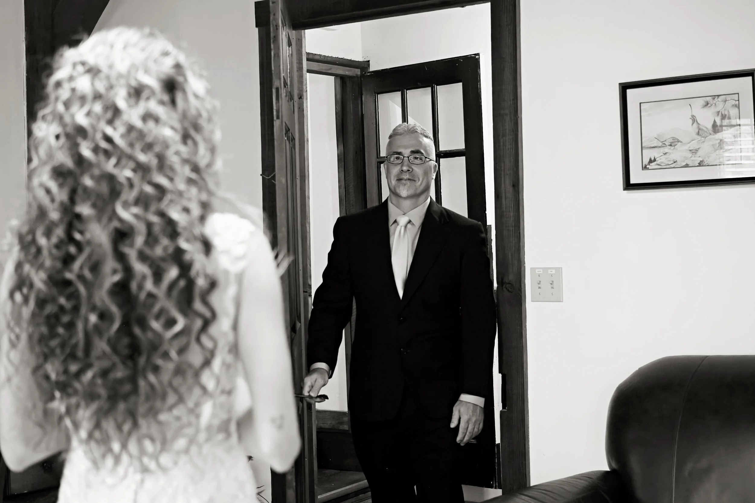 A man in a suit and glasses standing in a doorway, with a woman with curly hair, wearing a patterned top, partially visible in the foreground, inside a room with framed pictures on the wall and dark wooden doors.