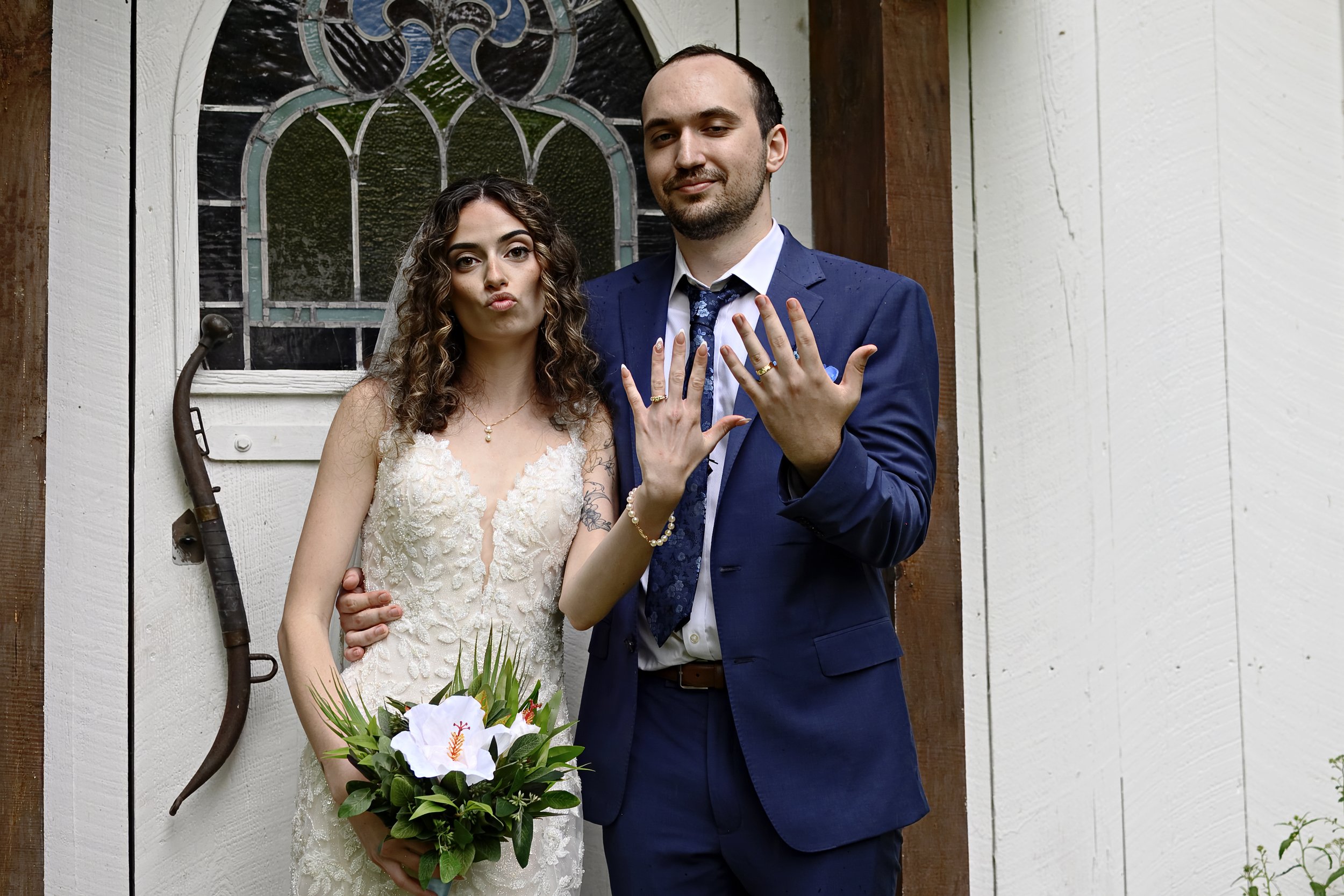 A newlywed couple stands in front of a stained glass window and white wooden wall; the woman is holding a bouquet and the man shows their rings on his hand.