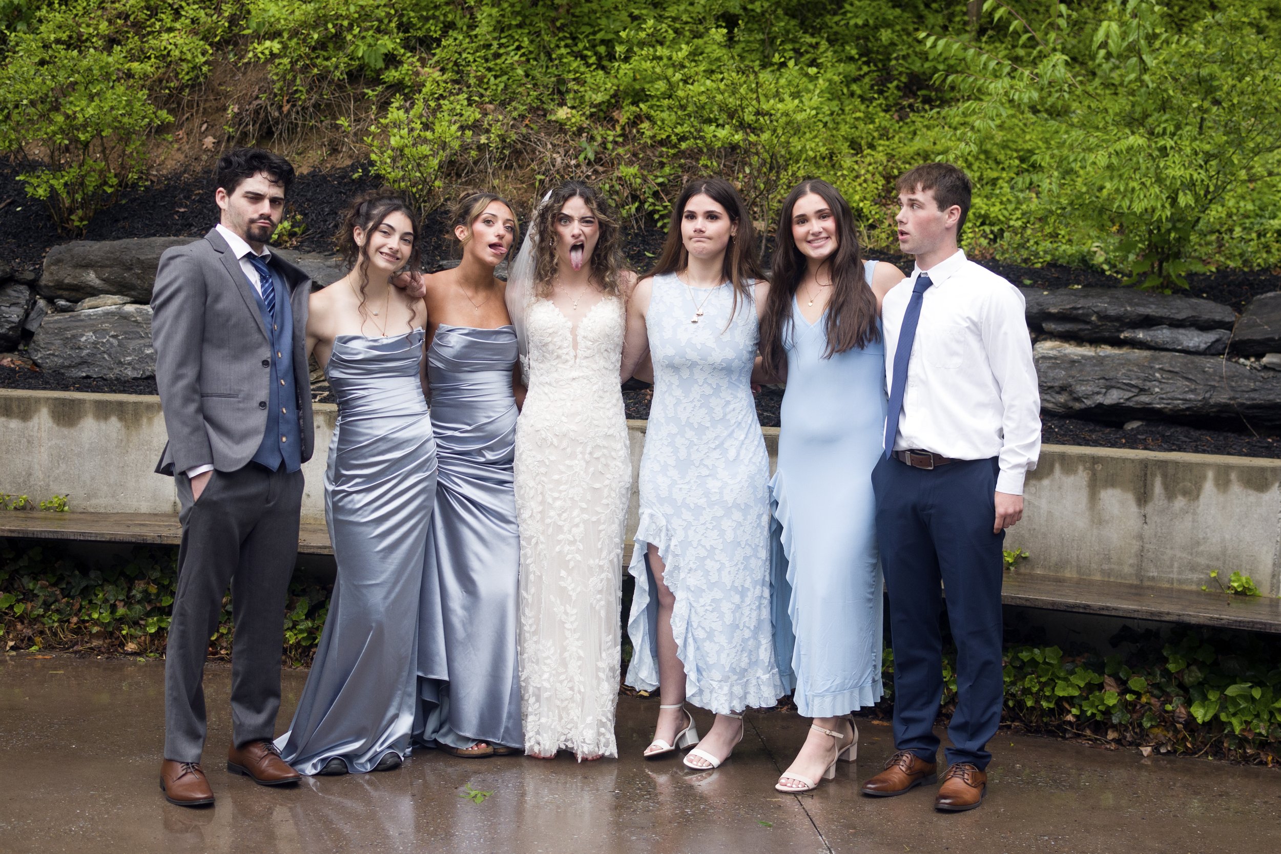 Group of seven young adults standing outdoors on a concrete pathway, with green bushes and rocks in the background. They are dressed in formal and semi-formal attire, with some making playful or silly facial expressions.