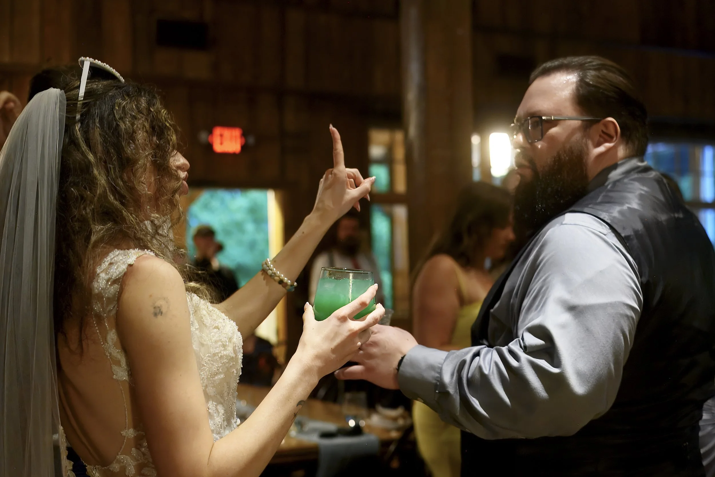 A woman in a wedding dress holding a green cocktail, raising her finger to a man with glasses and a beard, who is dressed semi-formally, inside a wooden venue during a wedding celebration.