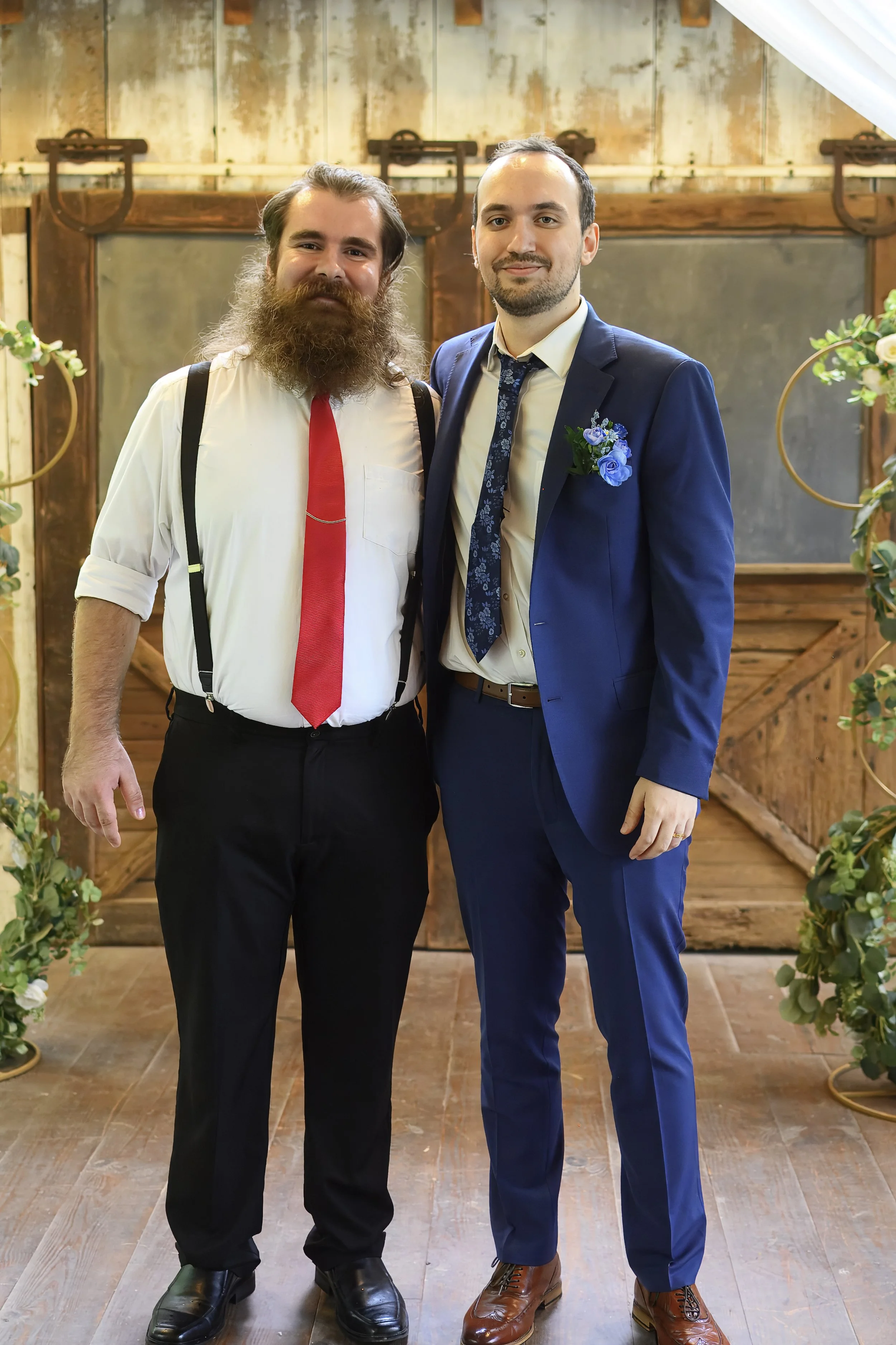 Two men dressed formally, standing side by side inside a rustic wooden building decorated with flowers and greenery, smiling at the camera.