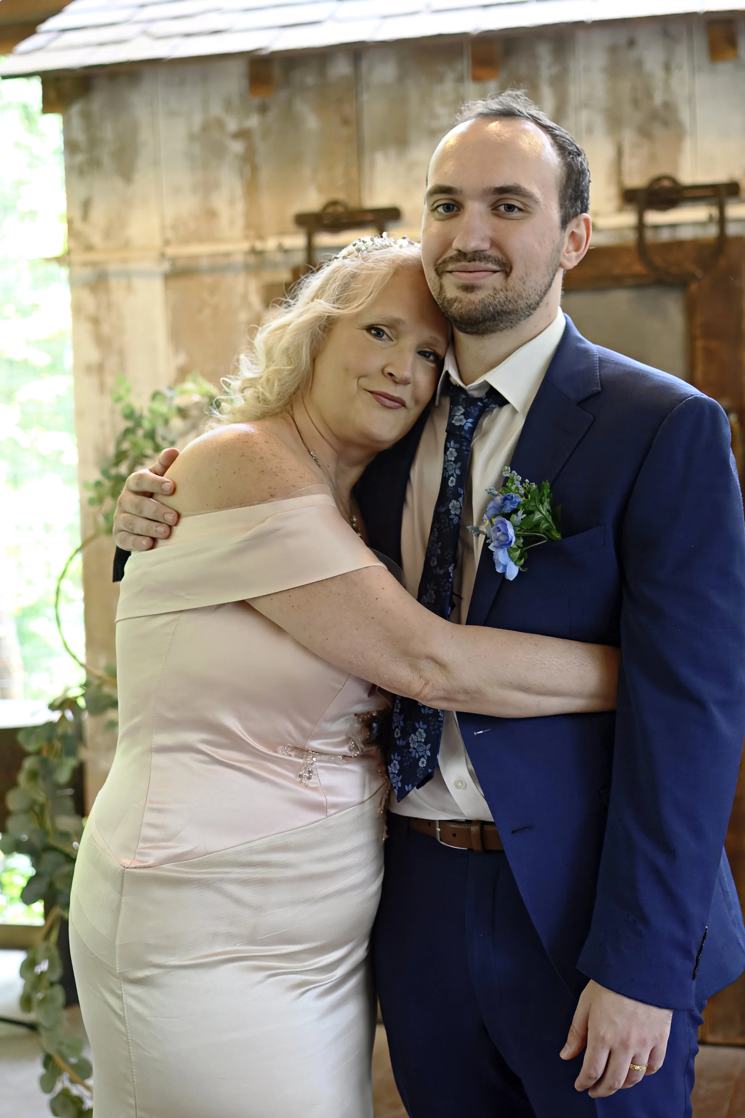 A woman and man hugging, dressed in formal attire, standing indoors with wooden walls and green plants in the background.