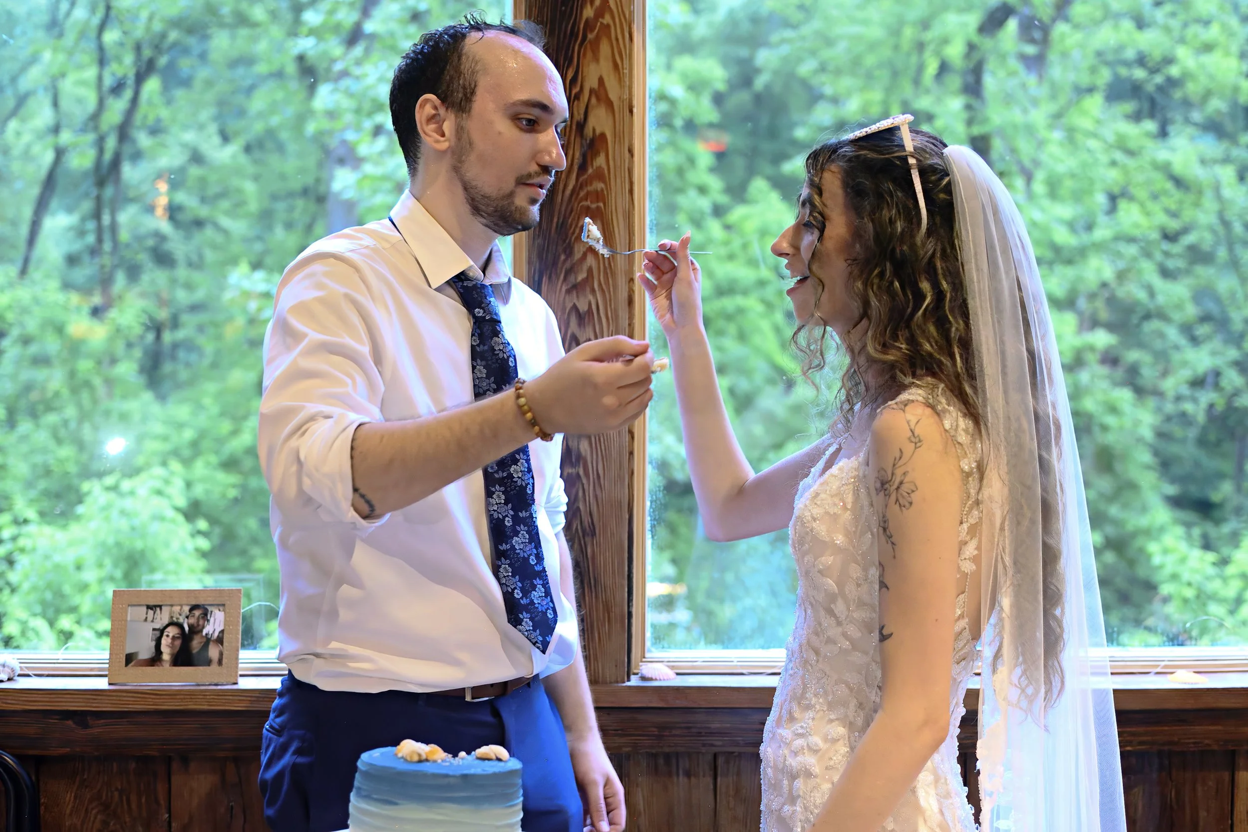 A man in a white shirt and dark tie feeding a piece of cake to a woman in a wedding dress with a veil, inside a wooden house with large windows and green trees outside.