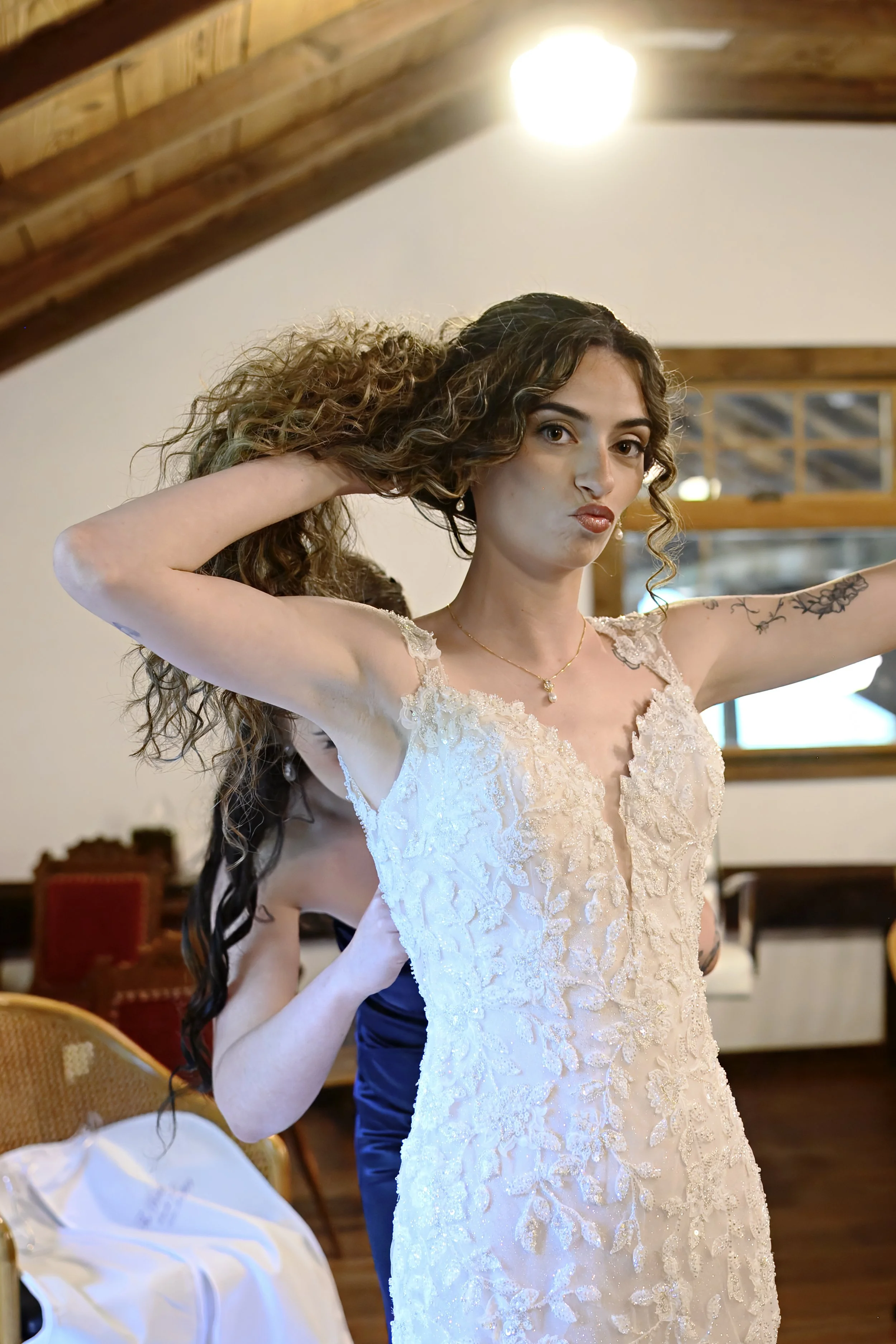 A woman in a lace wedding dress with curly hair stands indoors, posing for a photo with a playful expression.