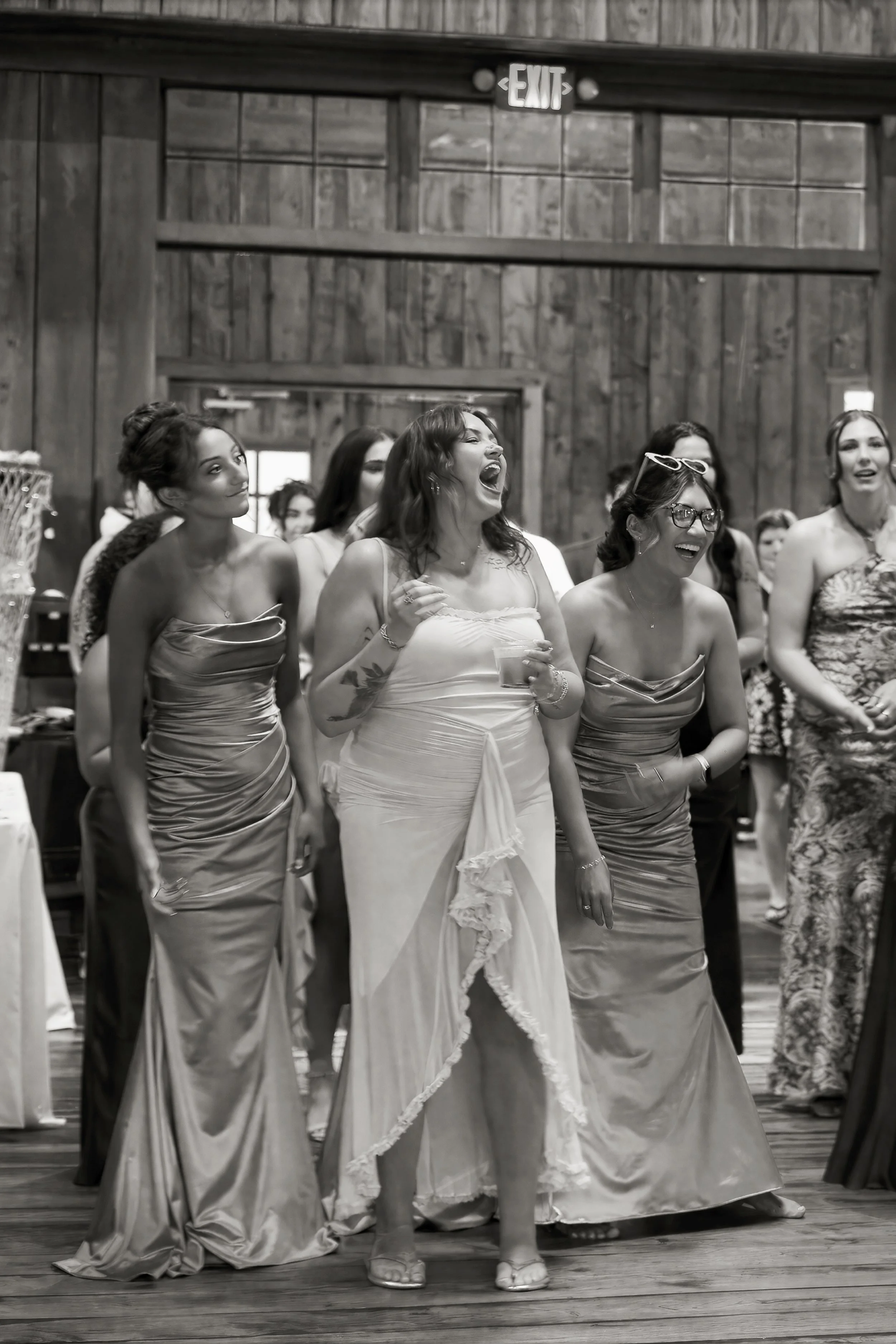 Group of women in formal dresses standing in a rustic wooden hall, some laughing and enjoying themselves.