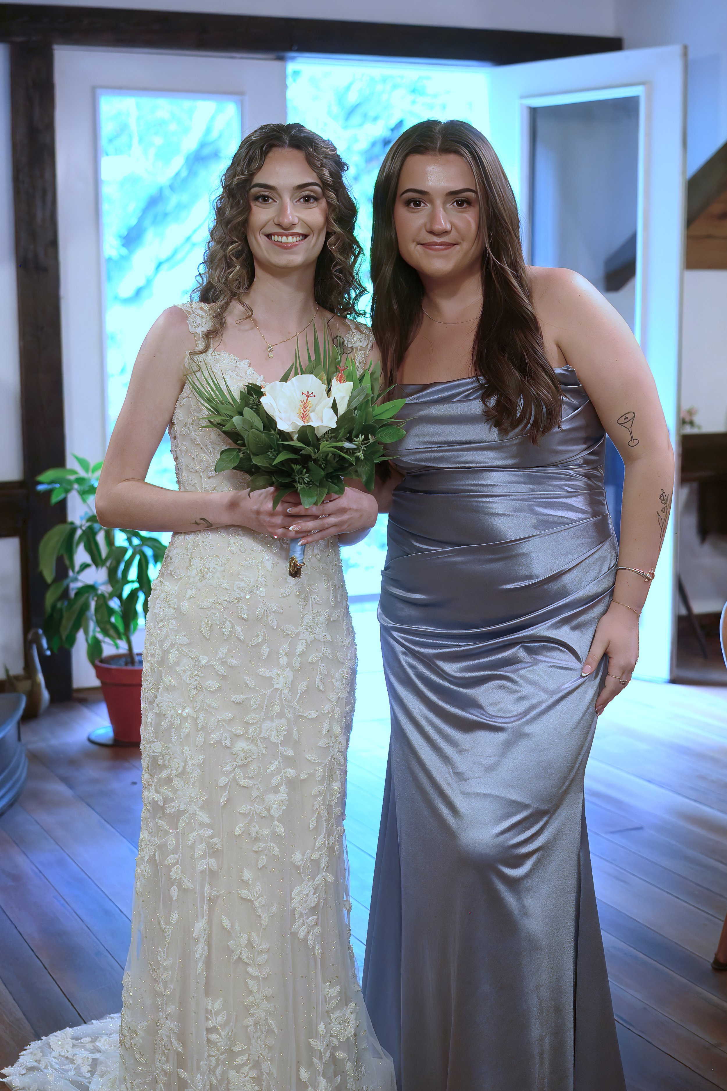 Two women standing indoors, one in a wedding dress holding a bouquet of white flowers, and the other in a silver satin dress, posing for a photo in front of a blue-lit background and potted plants.