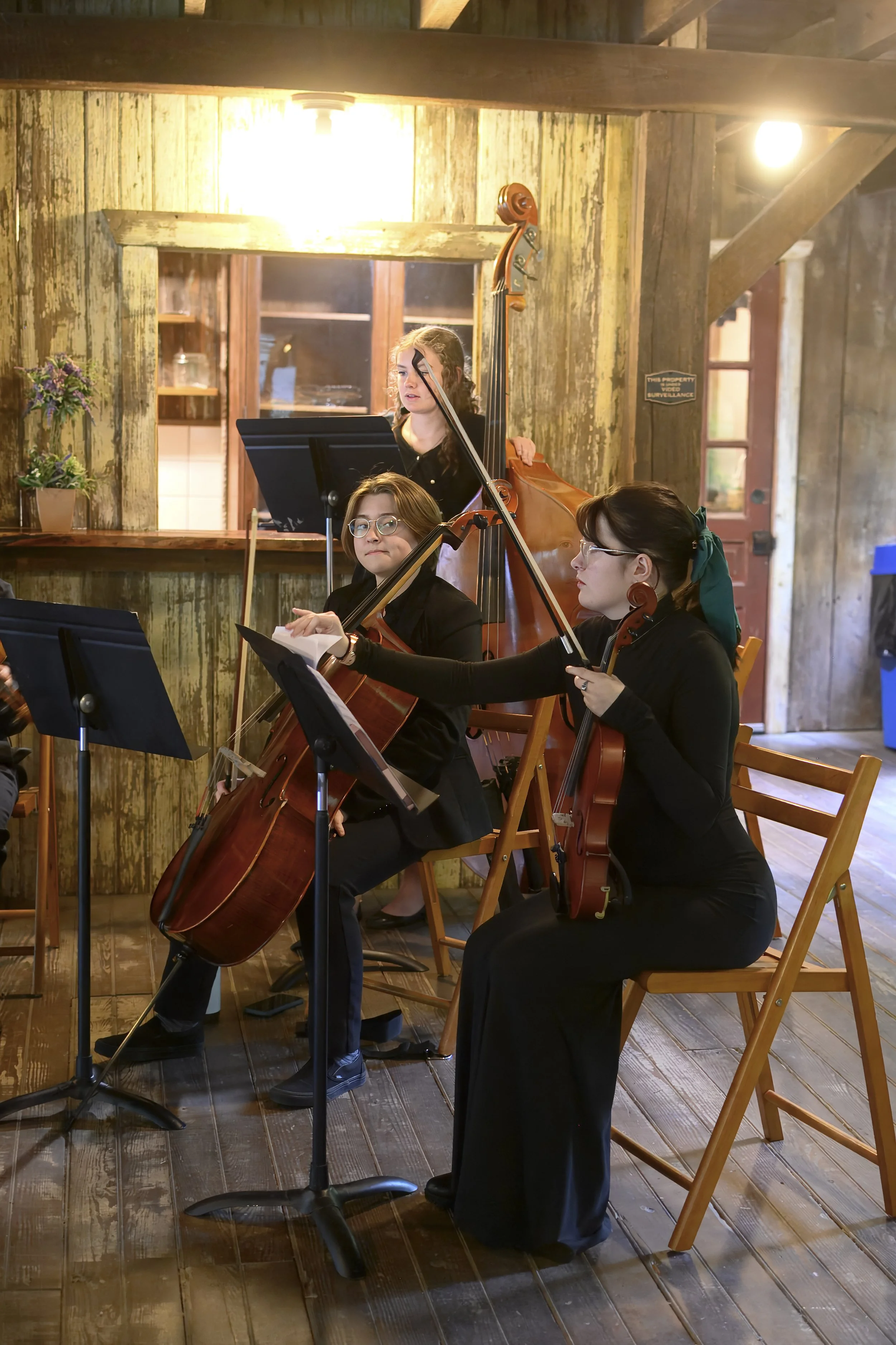 Three women musicians playing string instruments in an indoor rustic wooden setting. One woman is standing behind a music stand, the other two are seated, all wearing black clothing and glasses.