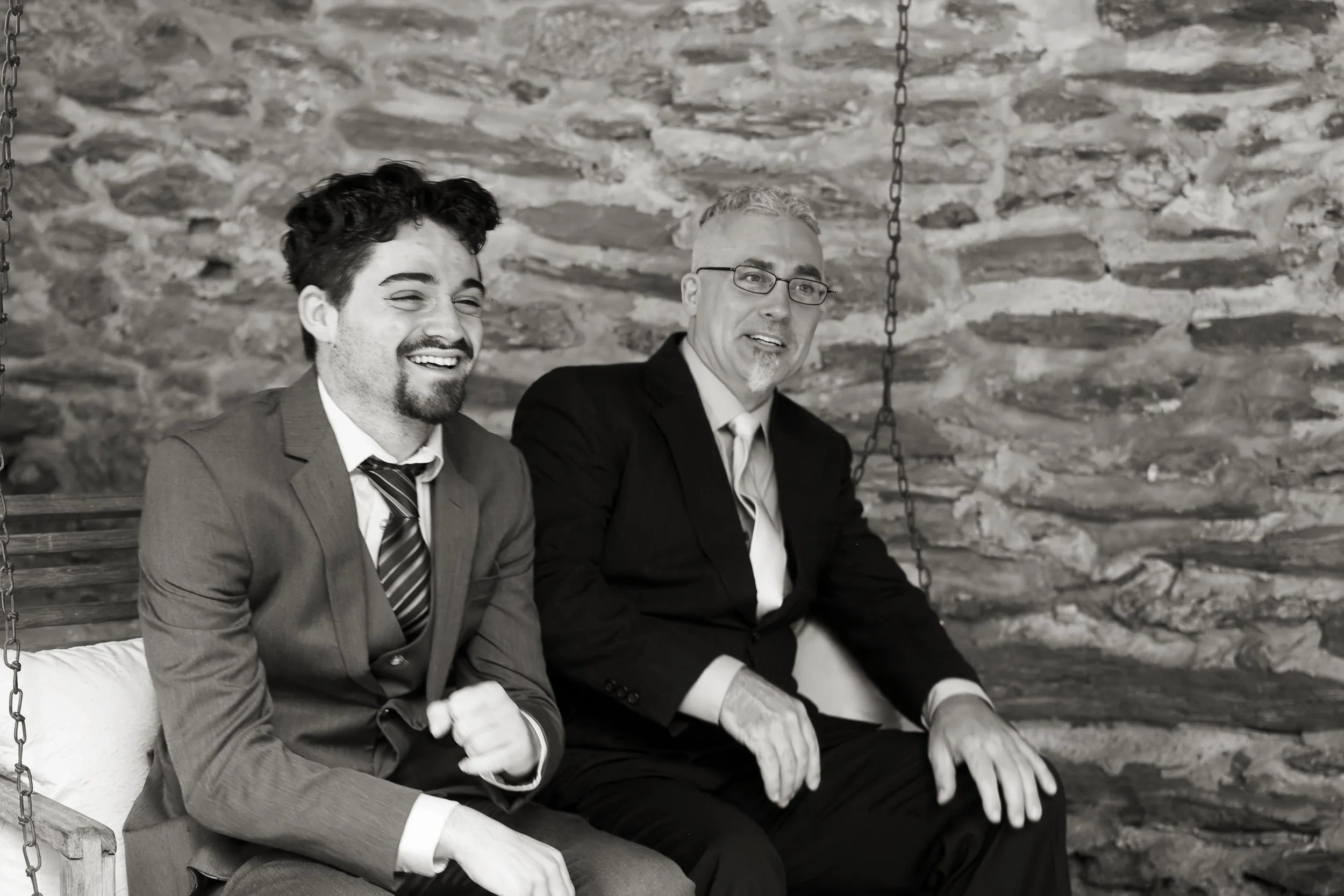 Two men in suits sitting on a wooden swing in front of a stone wall. The man on the left is smiling, wearing glasses and a tie. The man on the right is also smiling, wearing glasses and a white shirt with a dark jacket.