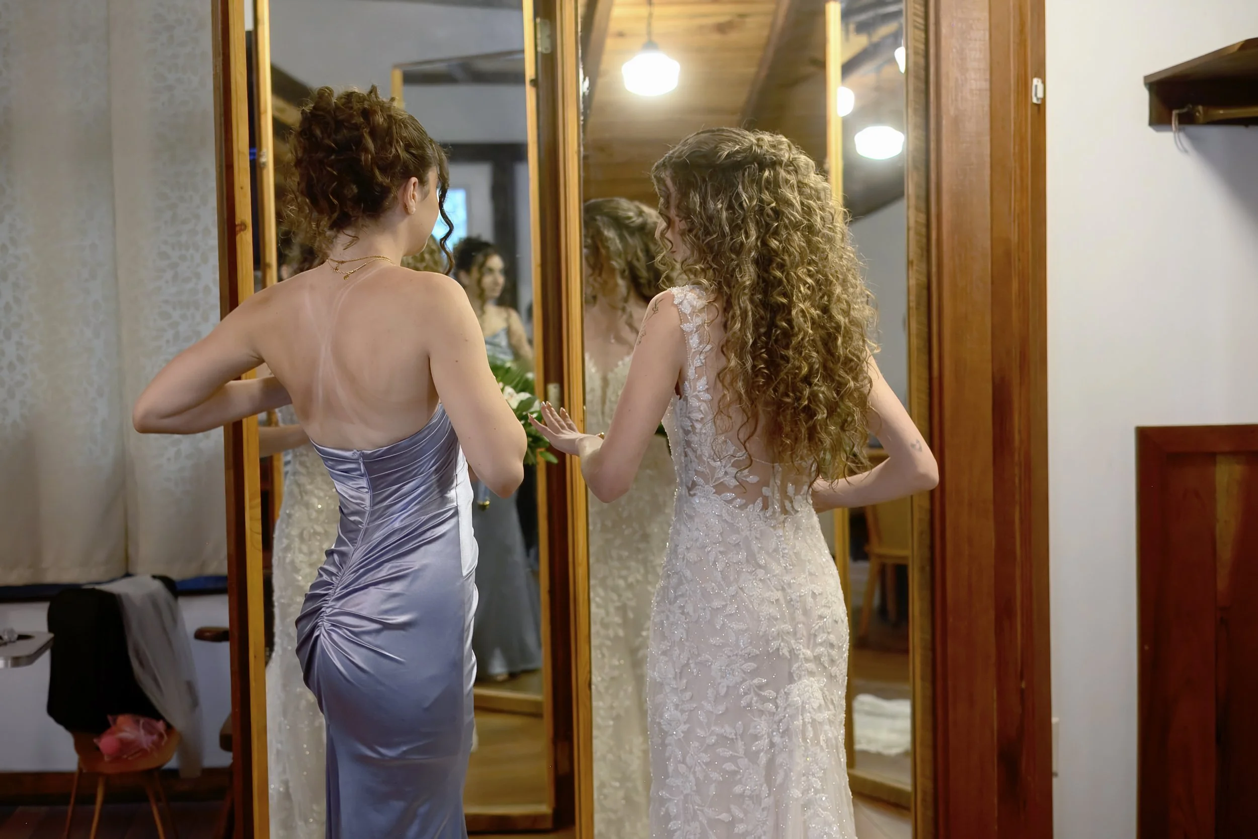 Two women, one in a lavender satin dress and the other in a white lace wedding gown, standing in front of a mirror, preparing for a wedding.