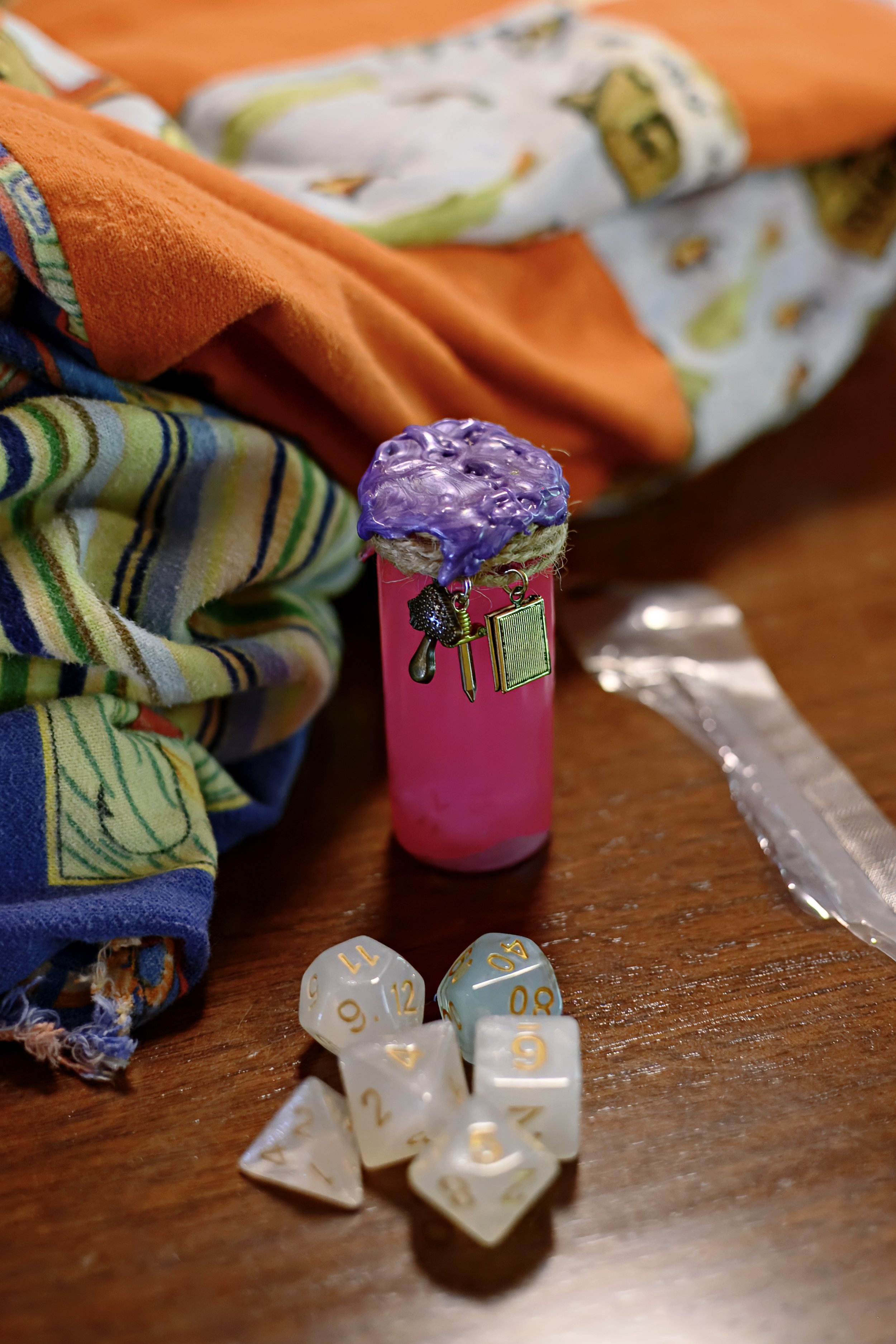 Close-up of a pink cylindrical object with purple-glazed baked item, small silver charms, and a sign hanging, with colorful cloths and a plastic ruler on a wooden surface in the background, and a scattering of white polyhedral dice in front.