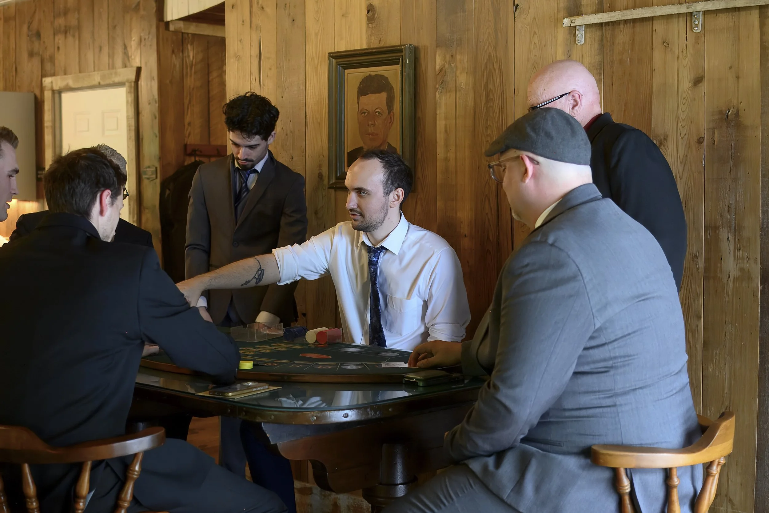 Men playing poker at a casino table in a room with wooden walls, with one seated man in a white shirt and blue tie, others standing or sitting nearby, and a portrait of a woman hanging on the wall.