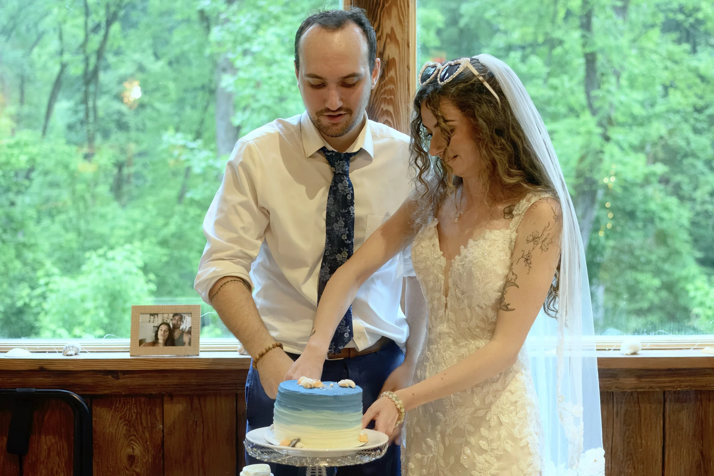 A bride and groom cutting their wedding cake together indoors near a large window with a view of green trees. A framed photo of two people is on the window sill in the background.