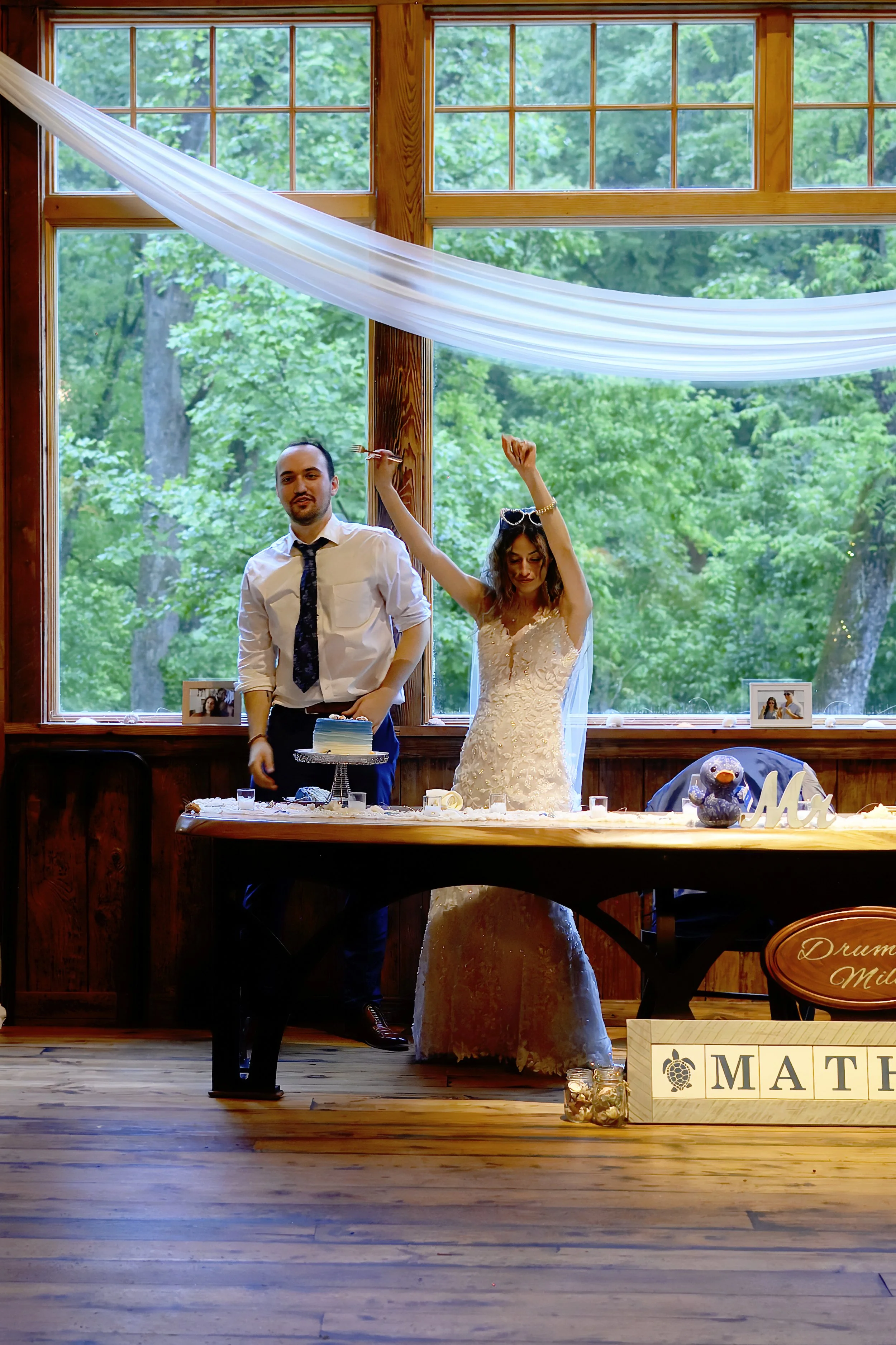 A newlywed couple celebrating at a wedding reception indoors in front of a large window with green trees outside. The bride is wearing a white wedding gown and raising her arms, while the groom stands next to her with a cake in front of them. There a