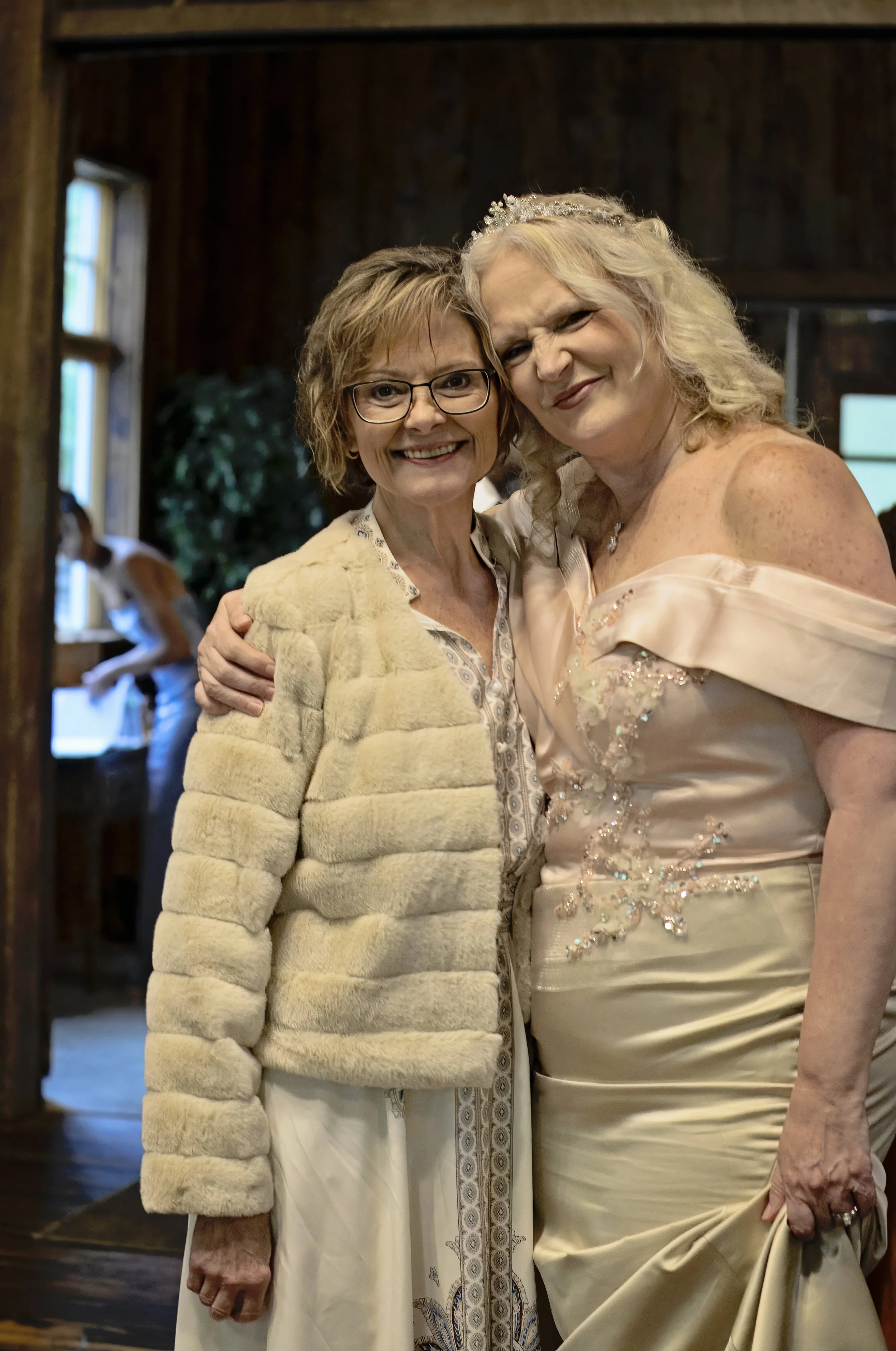 Two women are standing close together indoors, smiling at the camera. One woman is wearing glasses and a beige furry jacket, and the other is dressed in an elegant, off-the-shoulder satin gown with embellishments and a tiara in her hair.