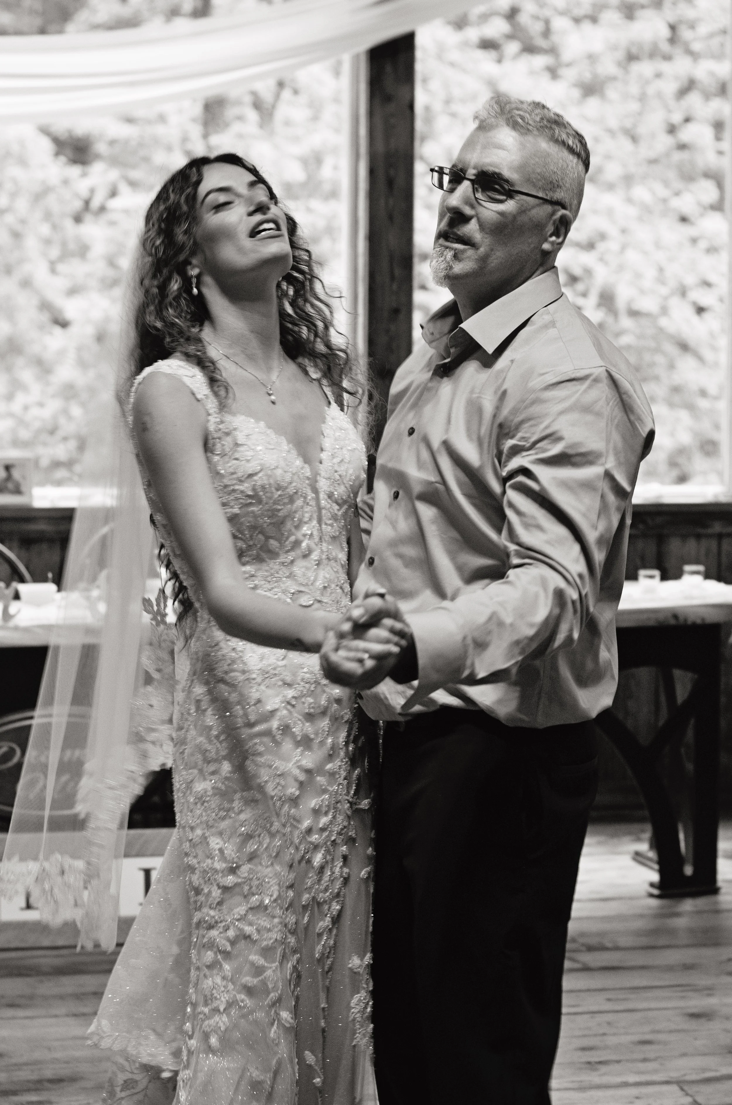 A black and white photo of a couple dancing at a wedding reception inside a rustic venue, with the bride in a lace wedding gown and the groom in a dress shirt and dark pants.