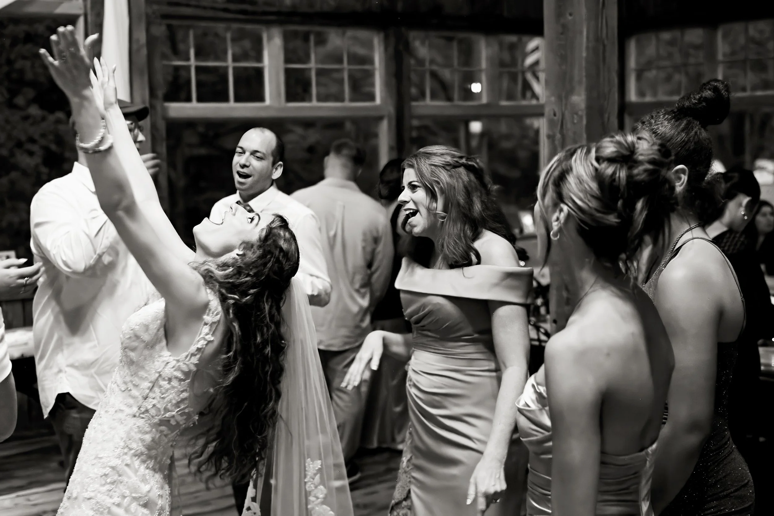 Couple dancing closely at a party, with a woman in a lace dress and a man in a white shirt, surrounded by other guests in a rustic interior