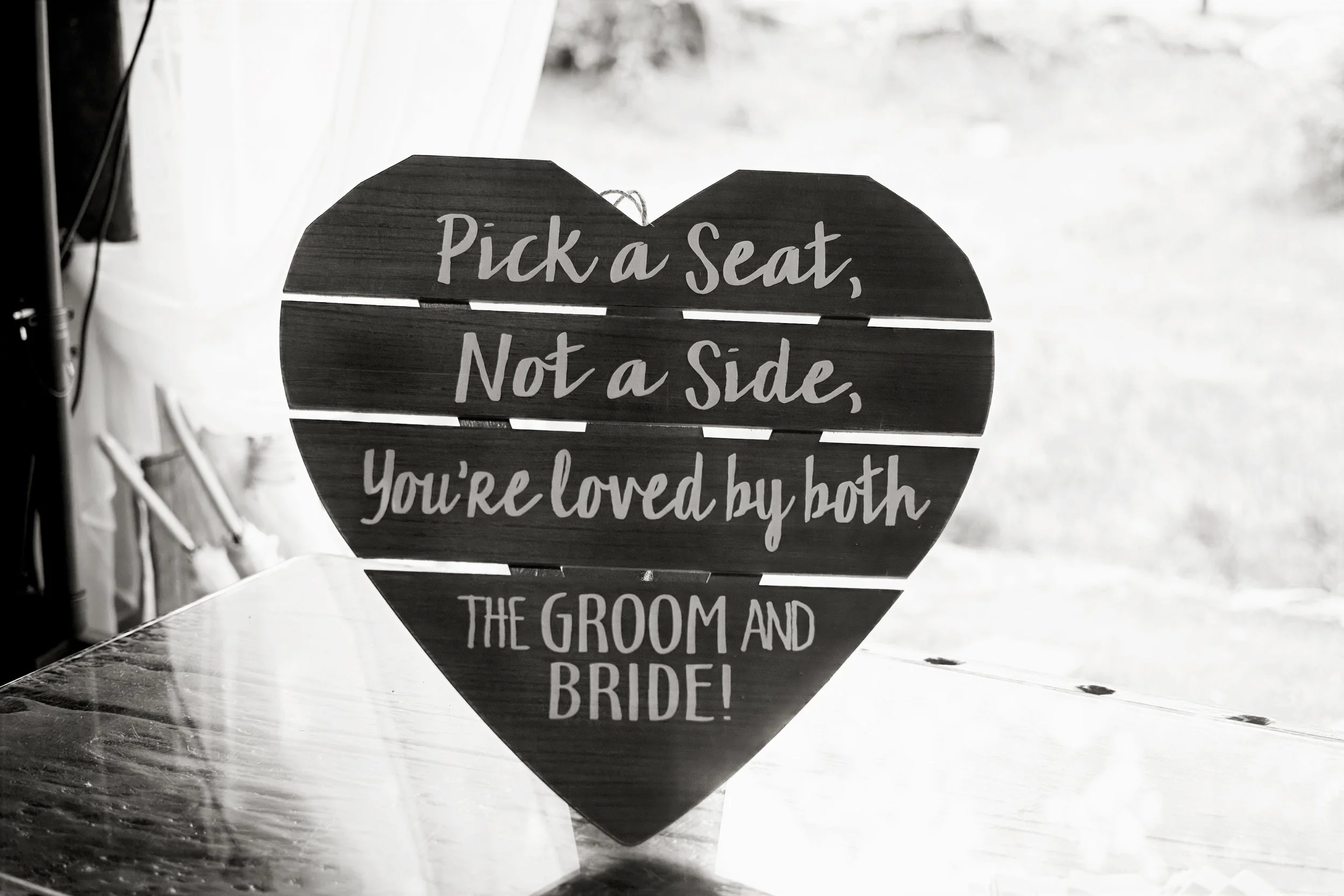 A heart-shaped wooden sign with white cursive text reading, 'Pick a seat, not a side, you're loved by both the groom and bride,' leaning against a wall on a wooden floor.