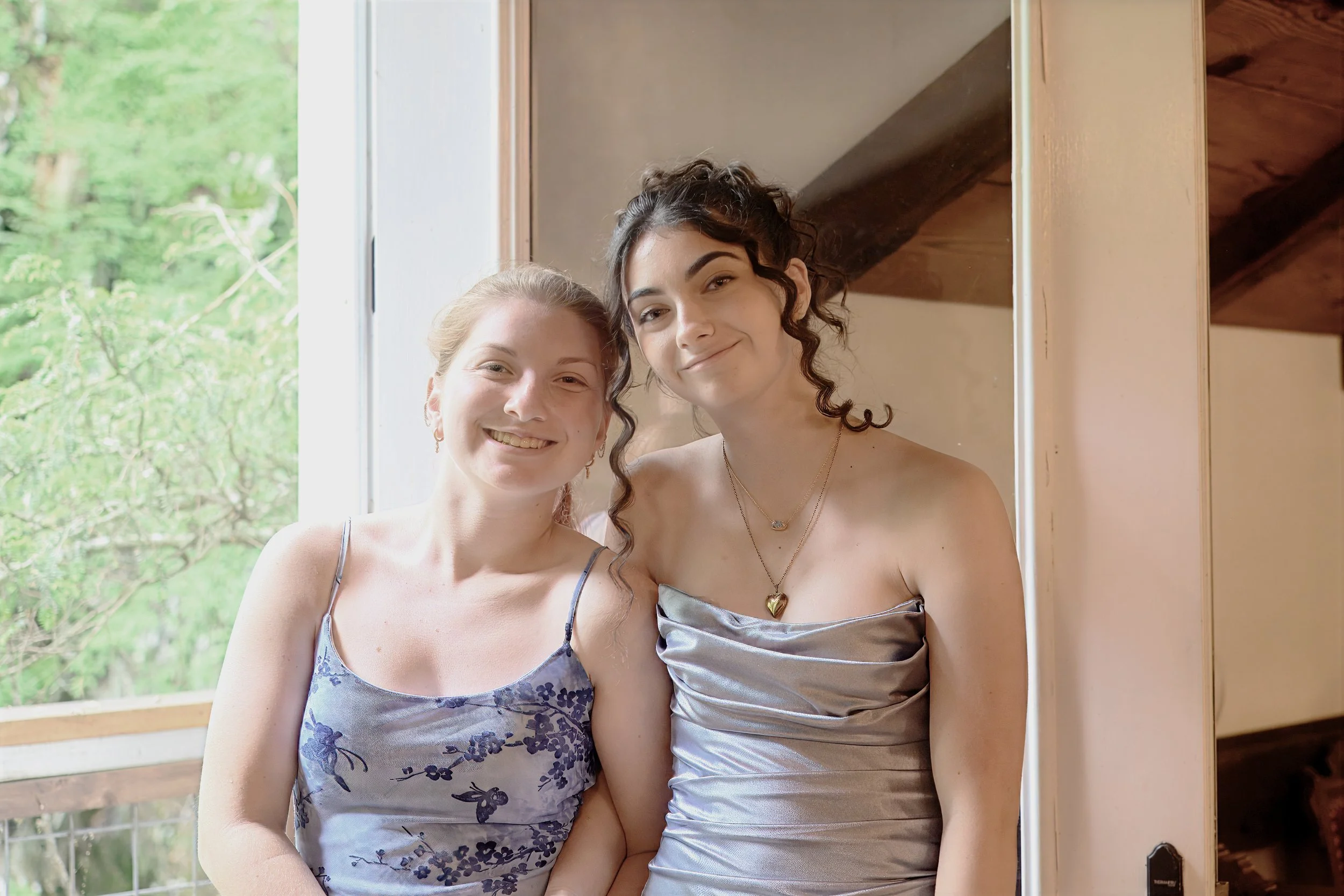 Two women smiling and posing together near a window, with a leafy green outdoor scene in the background.