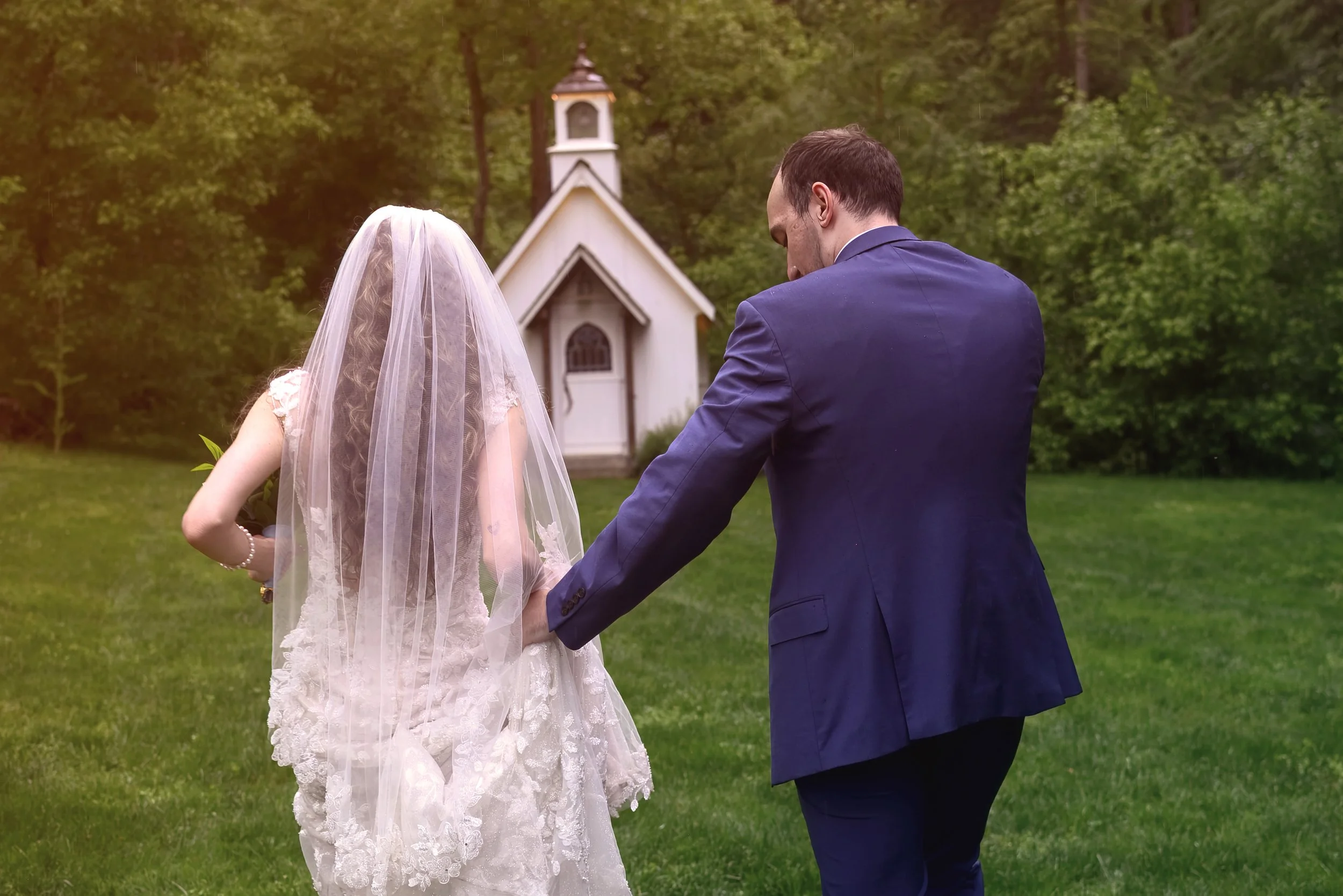 A bride and groom holding hands in a grassy outdoor area with trees and a small white chapel in the background.