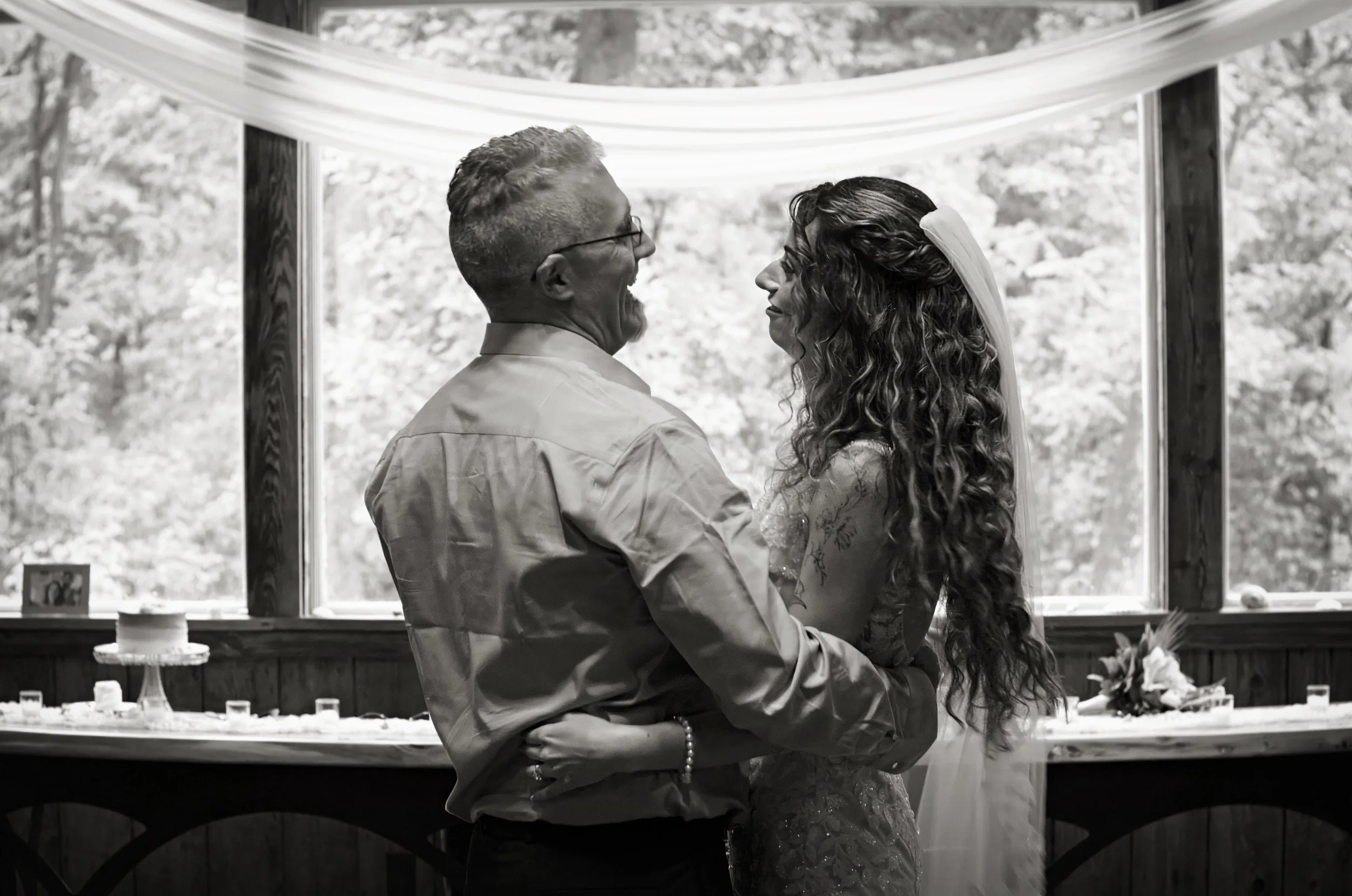 A couple dancing closely at a wedding reception with a window and outdoor trees in the background.