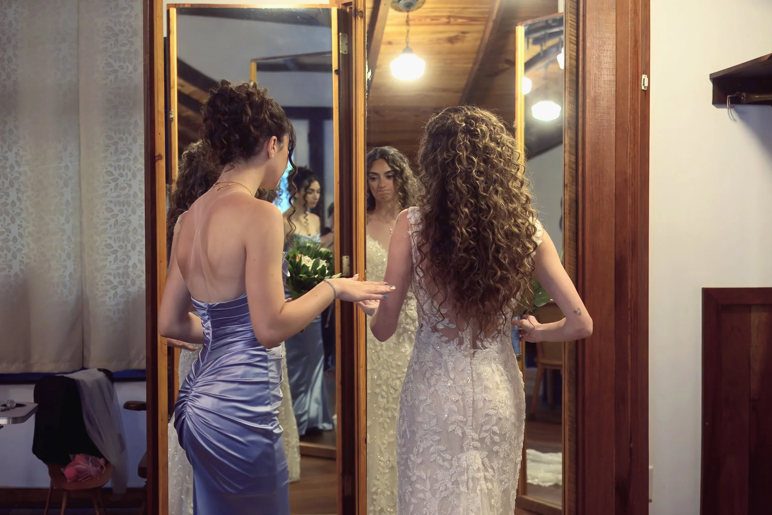 A bride with long, curly hair wearing a lace wedding gown stares at her reflection in a mirror, while a bridesmaid in a shiny, light purple dress stands nearby, helping her organize her appearance in a warmly lit room with wooden ceiling and decor.