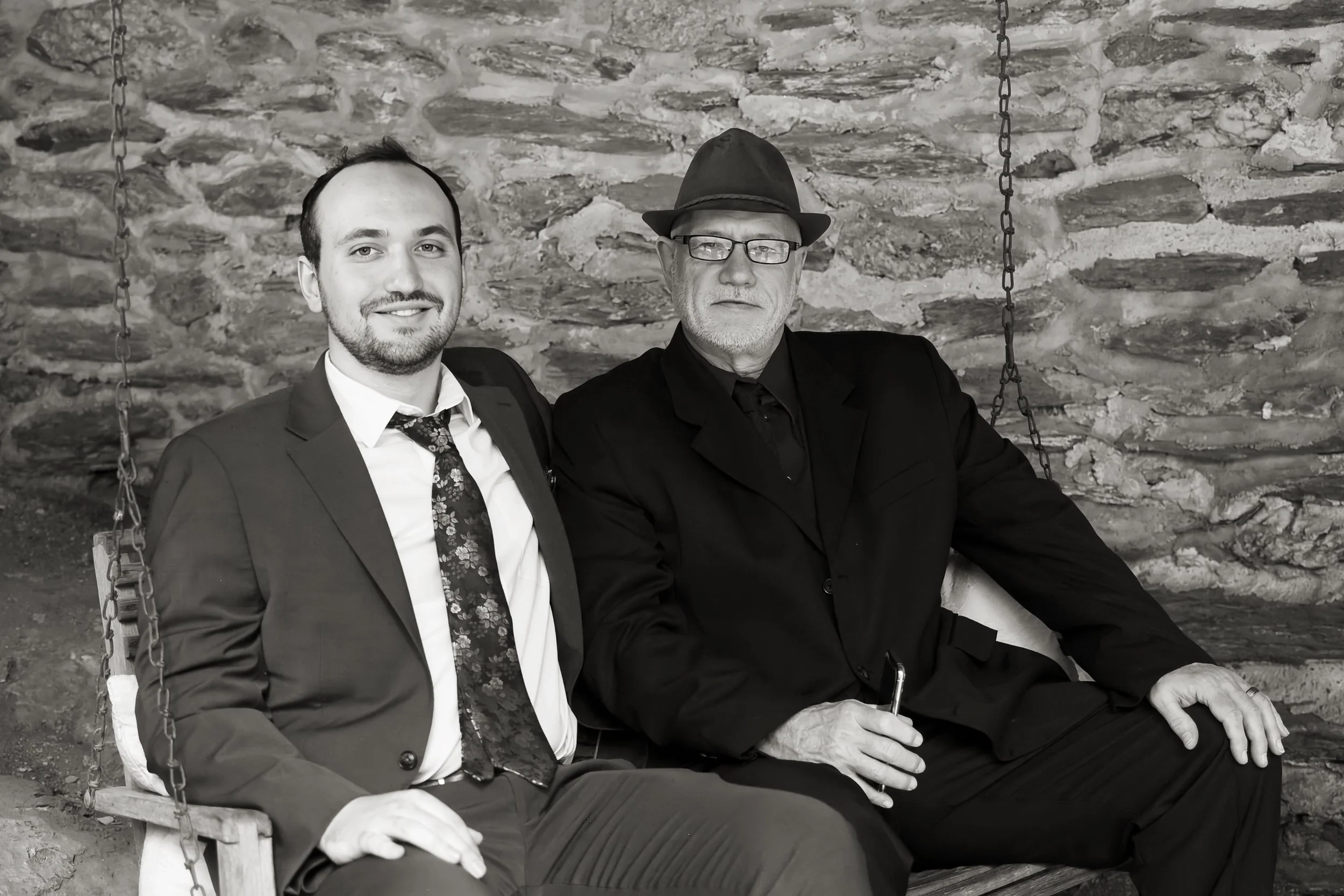 Two men sitting on a wooden swing with metal chains in front of a stone wall. The man on the left is smiling, wearing a suit, white shirt, and a patterned tie. The man on the right has glasses, a beard, a dark hat, and dark clothing.