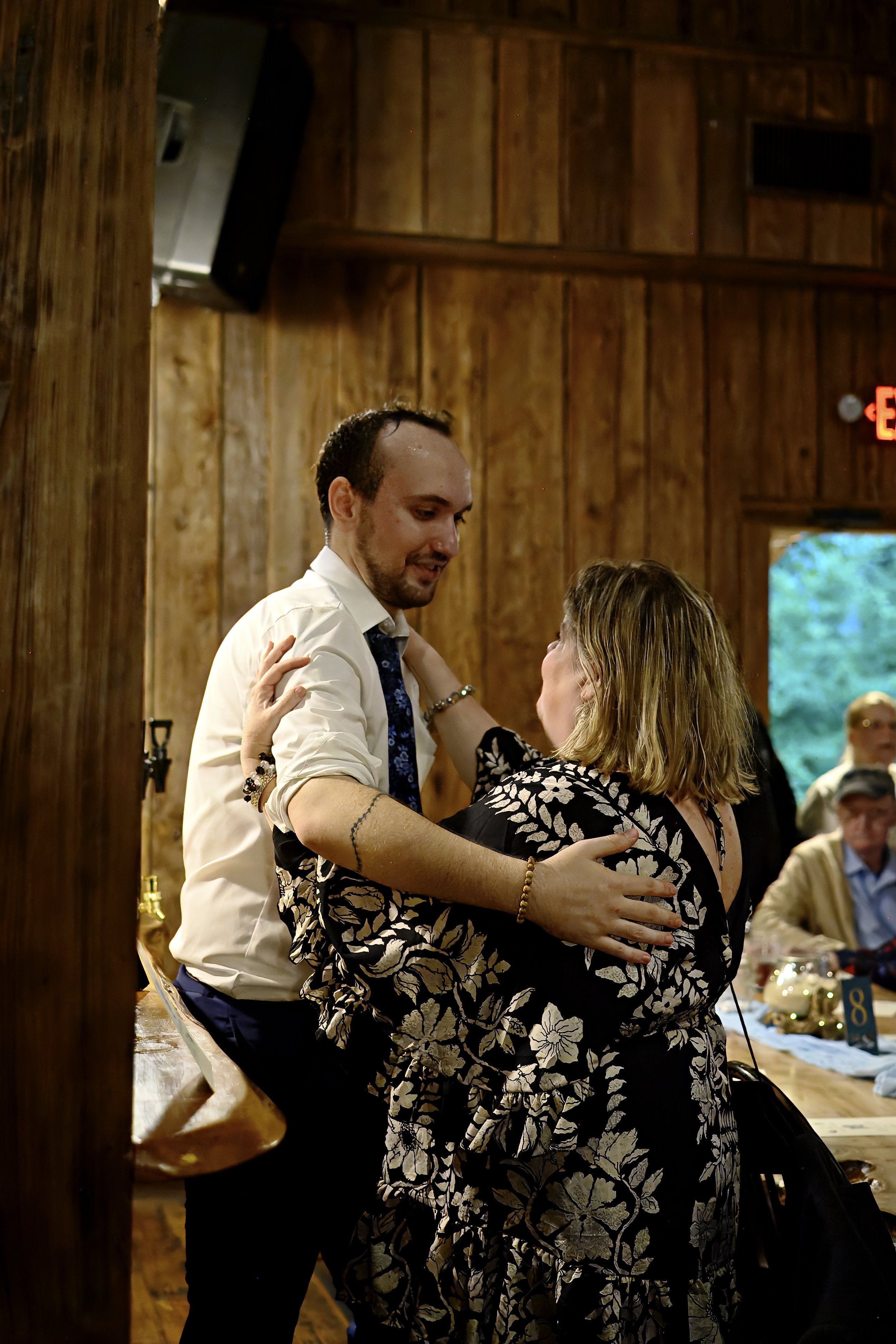 A man and woman dancing together at a wooden event space, with other people sitting at tables in the background.