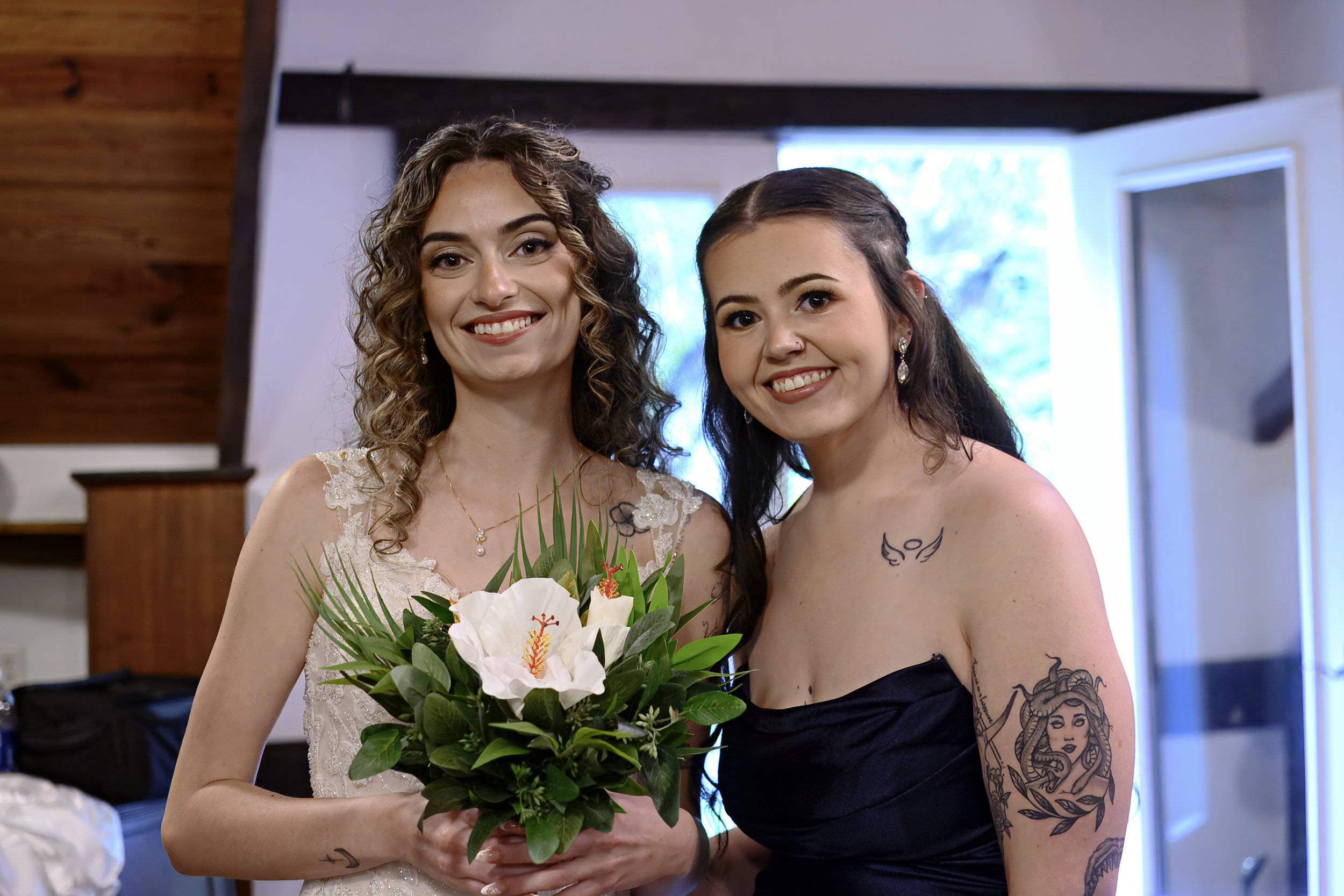 Two women smiling and taking a selfie indoors, one holding a bouquet of white lilies and green leaves, the other wearing a black dress with tattoos on her arm, both looking at the camera.